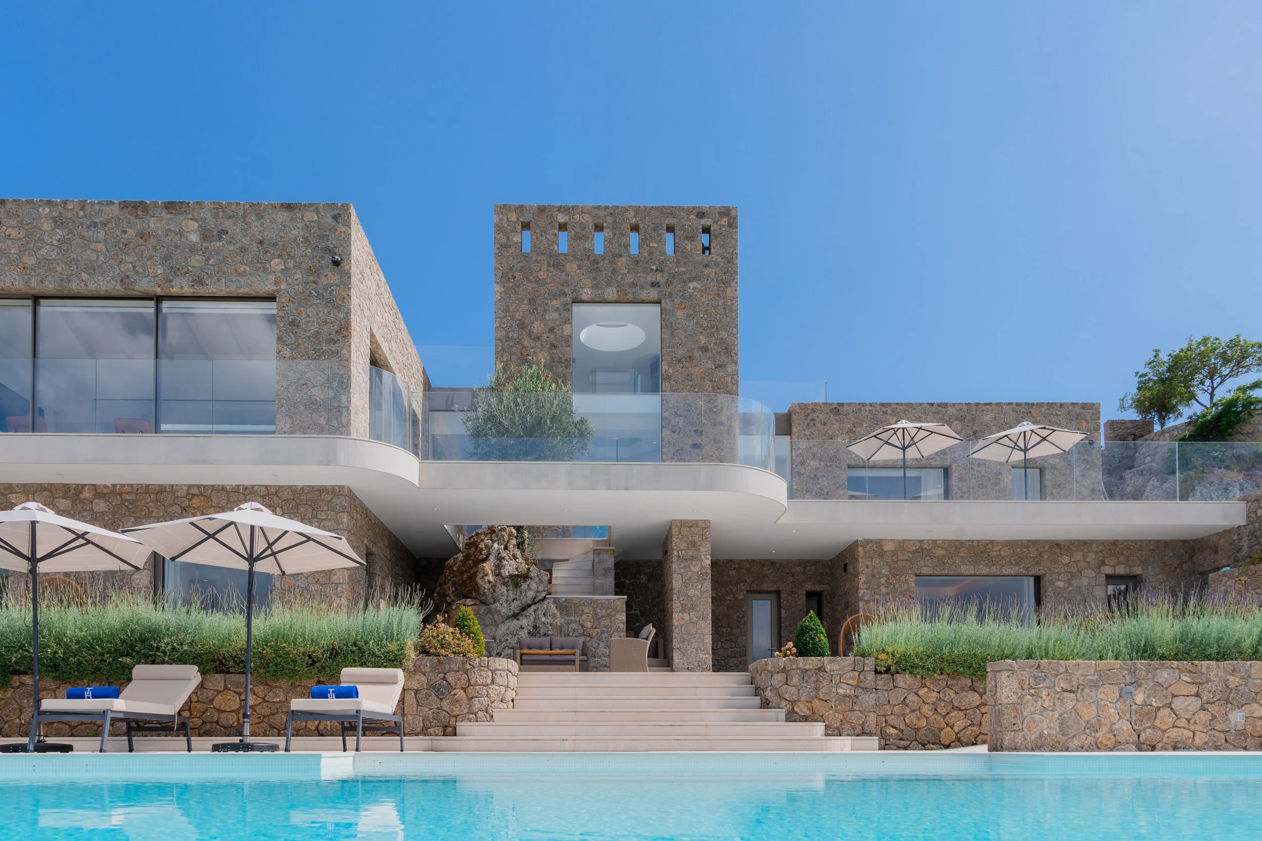 Luxury modern house with stone exterior, multiple levels, outdoor pool, lounge chairs, umbrellas, and greenery under a clear blue sky.