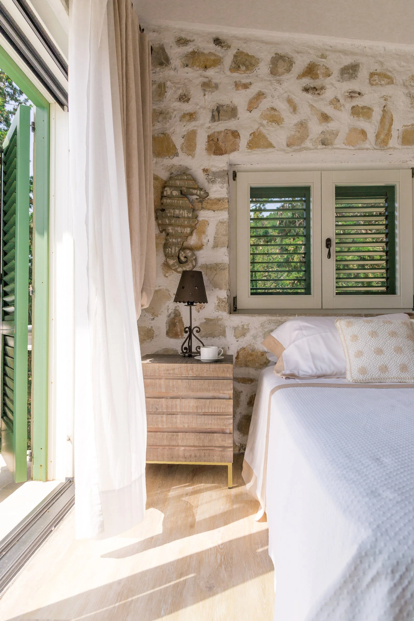 Sunlit bedroom with stone wall, green shutters on window, beige curtains, wooden nightstand, black lamp, coffee cup, and bed with white bedding and beige pillows.