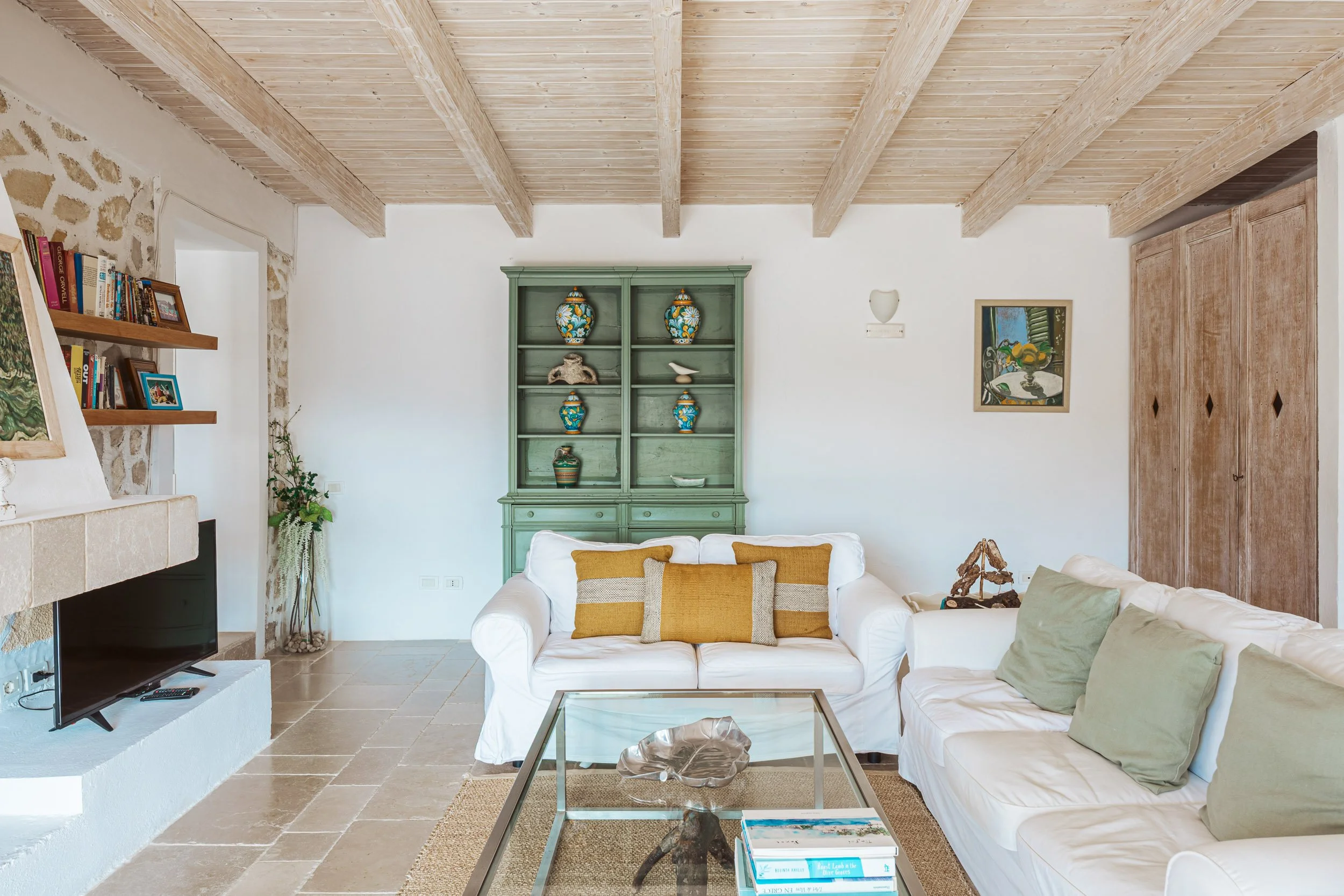 Living room with white couch and yellow throw pillows, glass coffee table with decorative bowl and books, green bookshelf with ceramics, flat-screen TV on a white stand, stone fireplace, wooden ceiling beams, framed artwork on white walls, potted pla