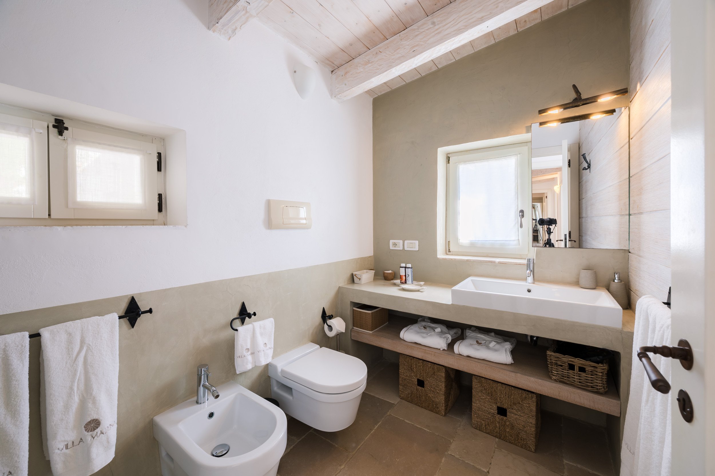 Bathroom with window, large mirror, white sink, toilet, bidet, beige and white walls, towel rack with white towels, wicker baskets, toiletries on counter, and a photographer's camera reflected in mirror.