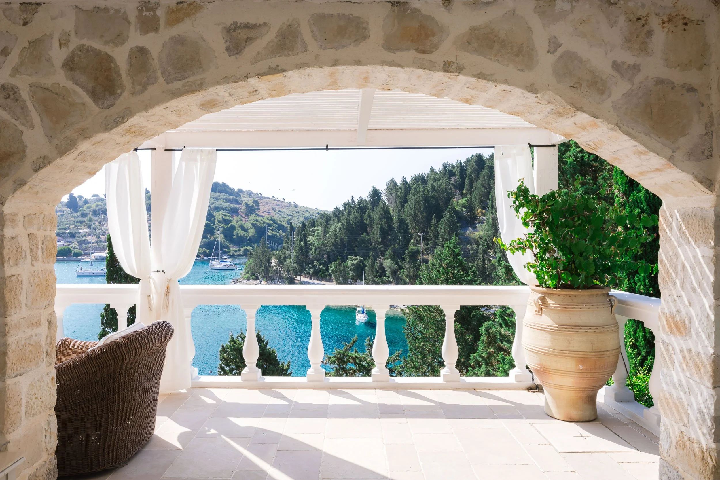 View of a balcony overlooking a scenic lake with sailboats, surrounded by lush green trees and hills, framed by a stone arch and white curtains.