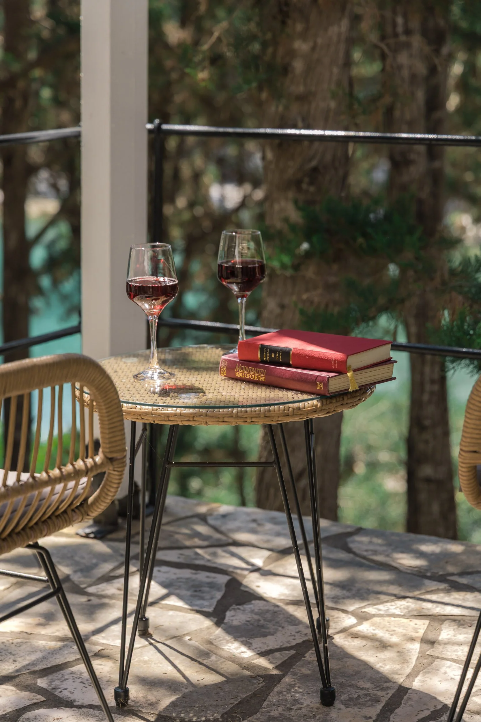 A round table with two glasses of red wine and three books on a balcony with trees in the background.