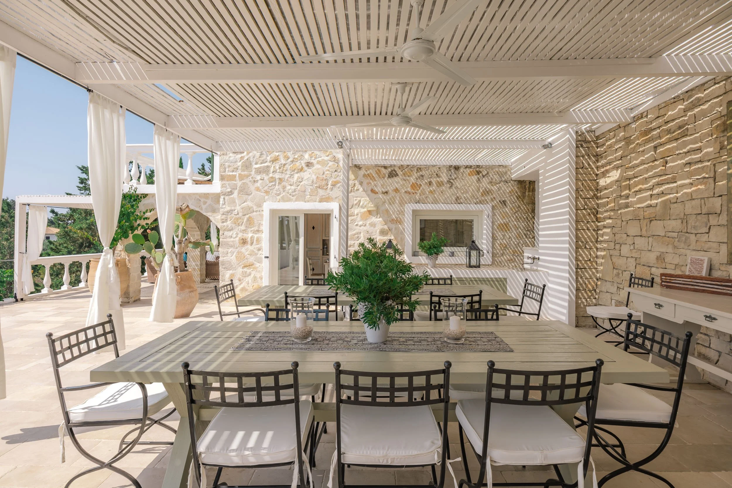 Outdoor patio with a large rectangular table, surrounded by black metal chairs with white cushions, decorated with a potted plant and candles, against a stone wall and white curtains on a sunny day.