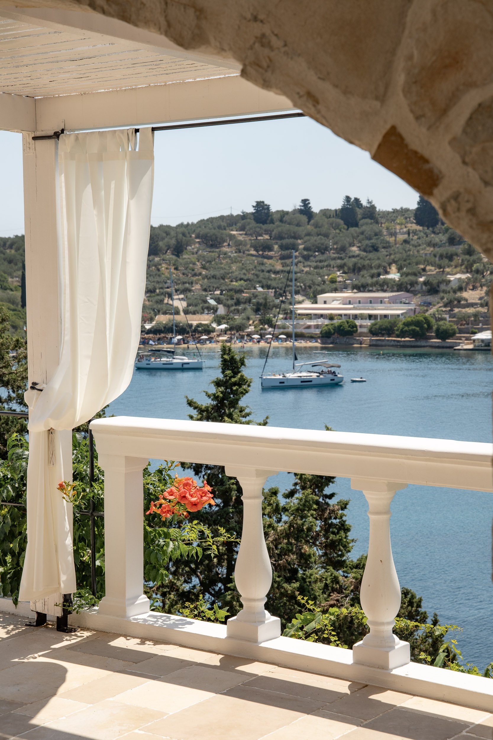 A view from a porch overlooking a river with sailboats, hills with houses and greenery in the background, and a white railing and curtain in the foreground.