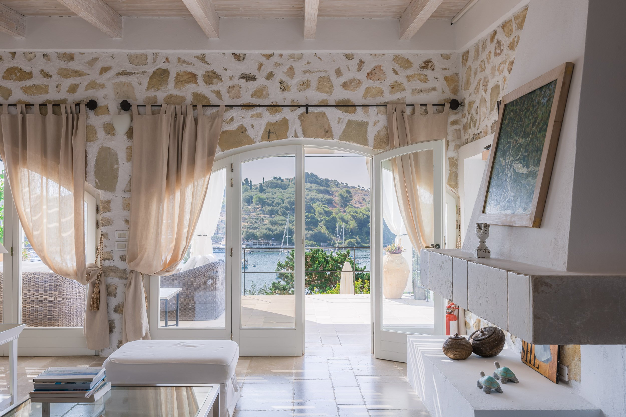 Living room with stone walls and large windows opening to a balcony view of a hillside and body of water.