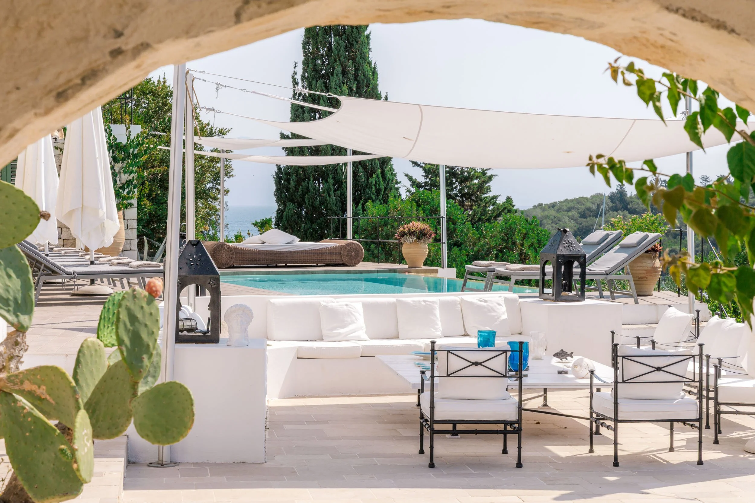 Outdoor pool area with white seating, lounge chairs, umbrellas, and greenery, overlooking the ocean in the background.