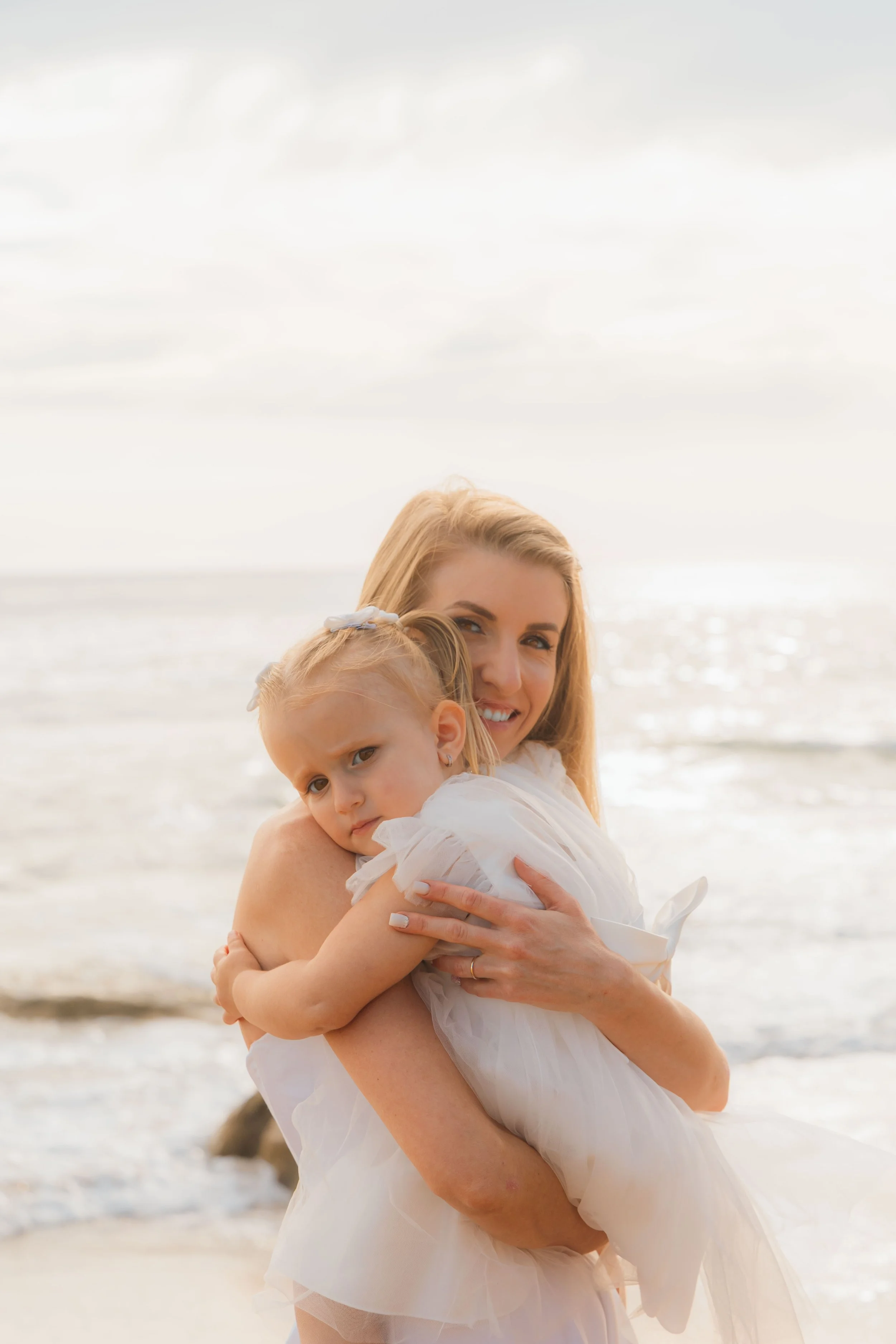 A woman holding a young girl on a beach with the ocean in the background.
