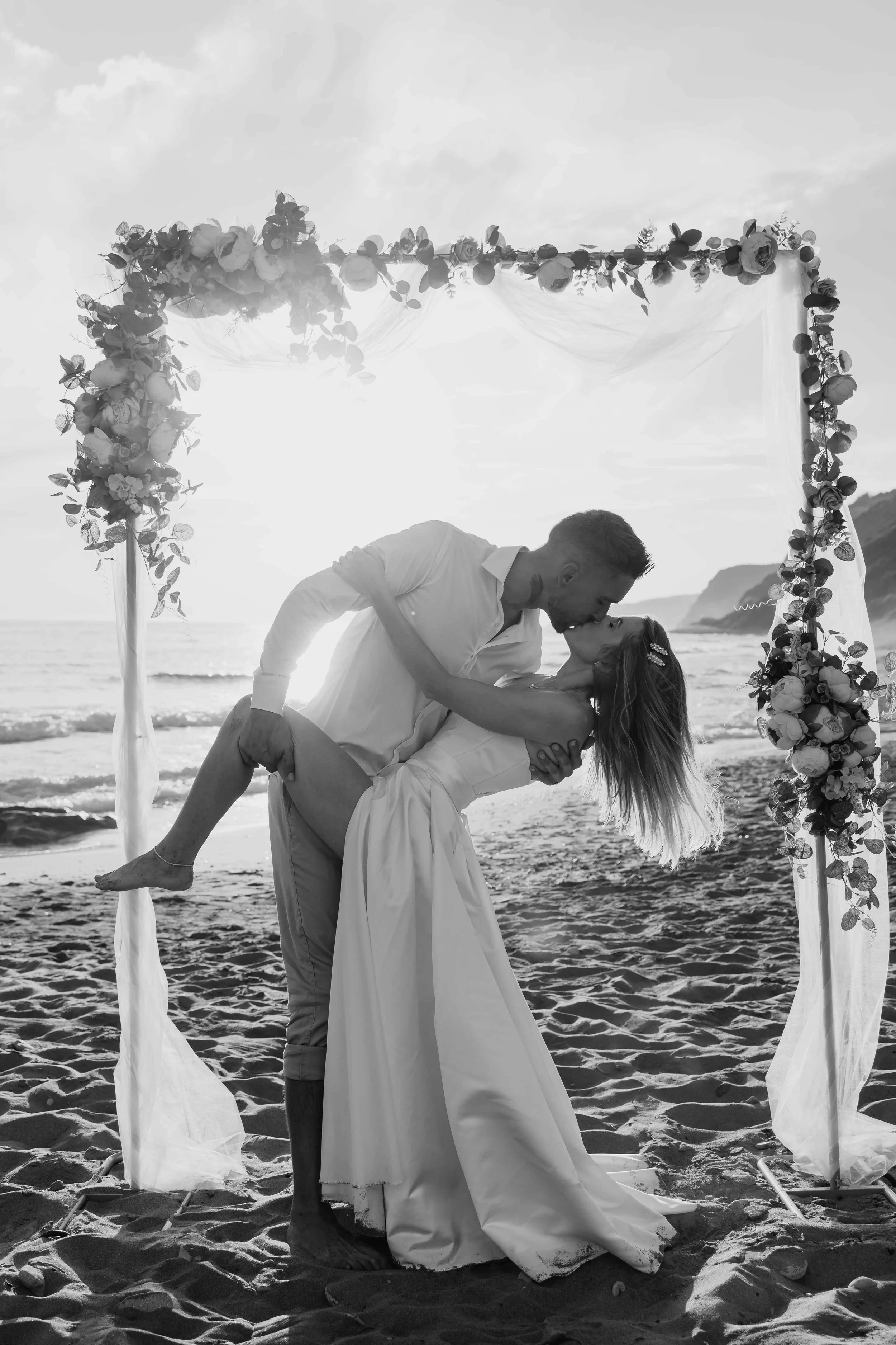 A couple is dancing on the beach under an arch decorated with flowers, with the ocean and a distant mountain in the background. The man is holding the woman, who is dipping backwards, and they are looking at each other lovingly.
