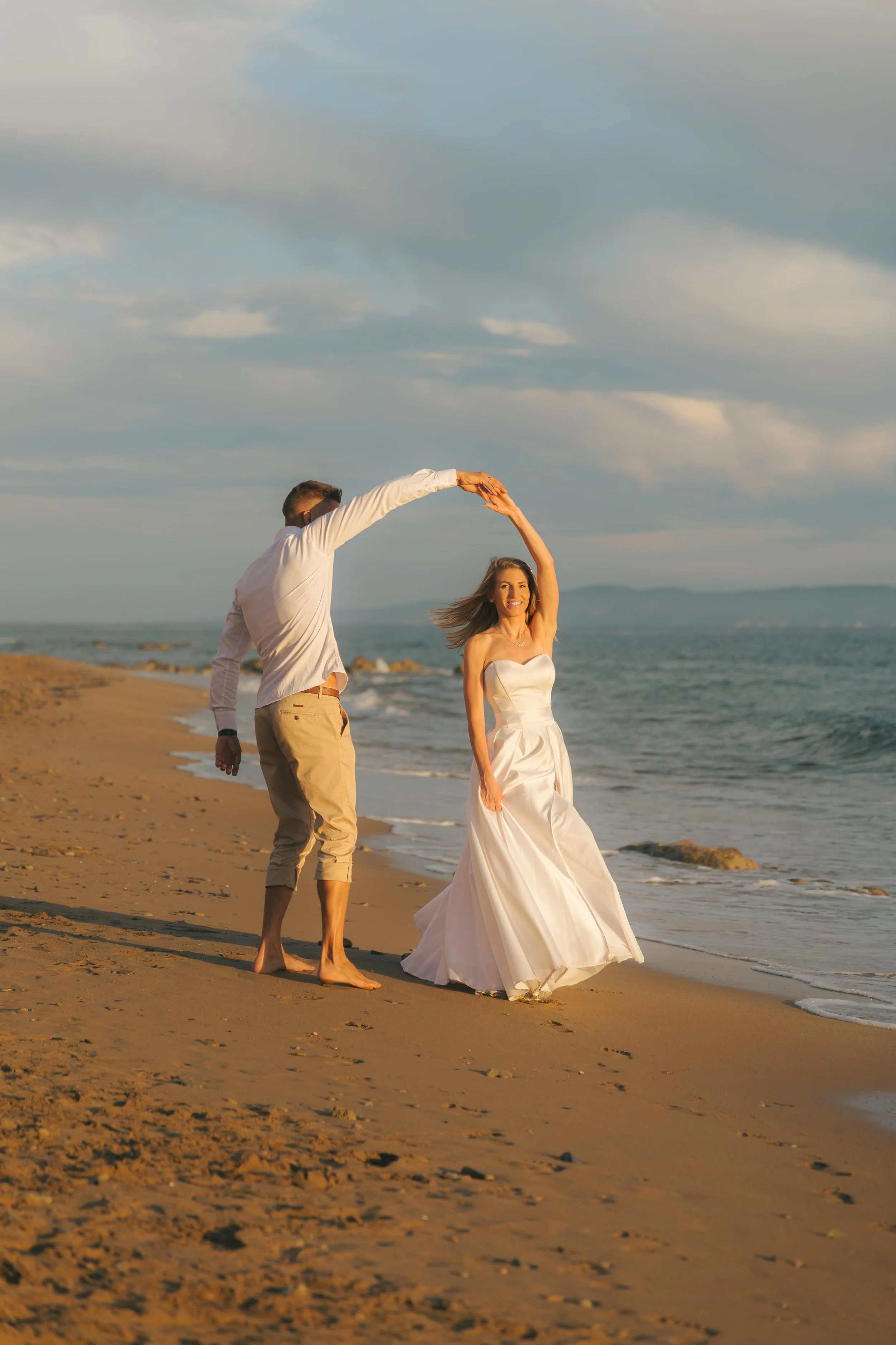 A couple dancing on the beach at sunset, with the man spinning the woman in a white dress, smiling, near the ocean waves.