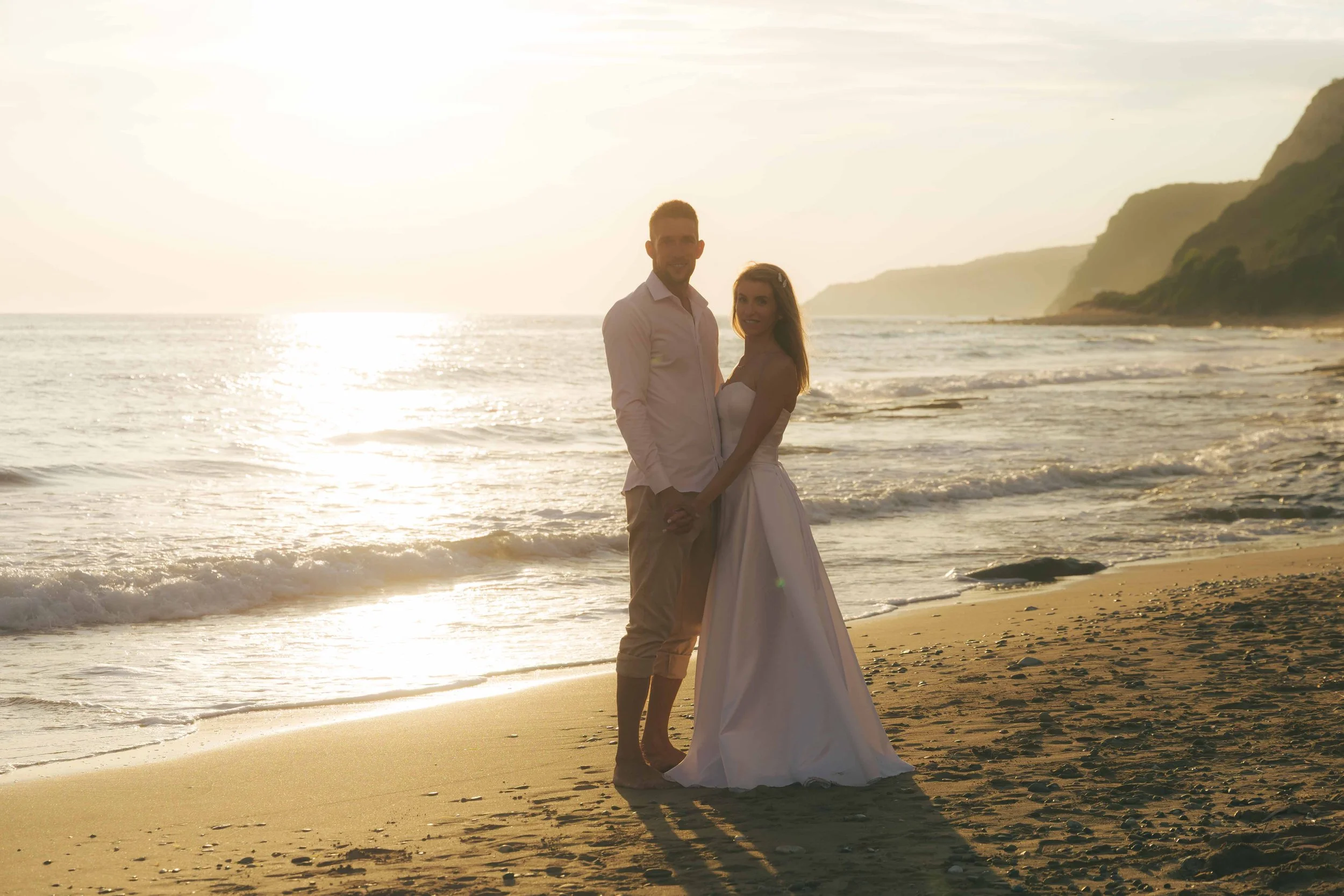 A couple dressed in white clothing standing hand in hand on a beach during sunset, with ocean waves and cliffs in the background.