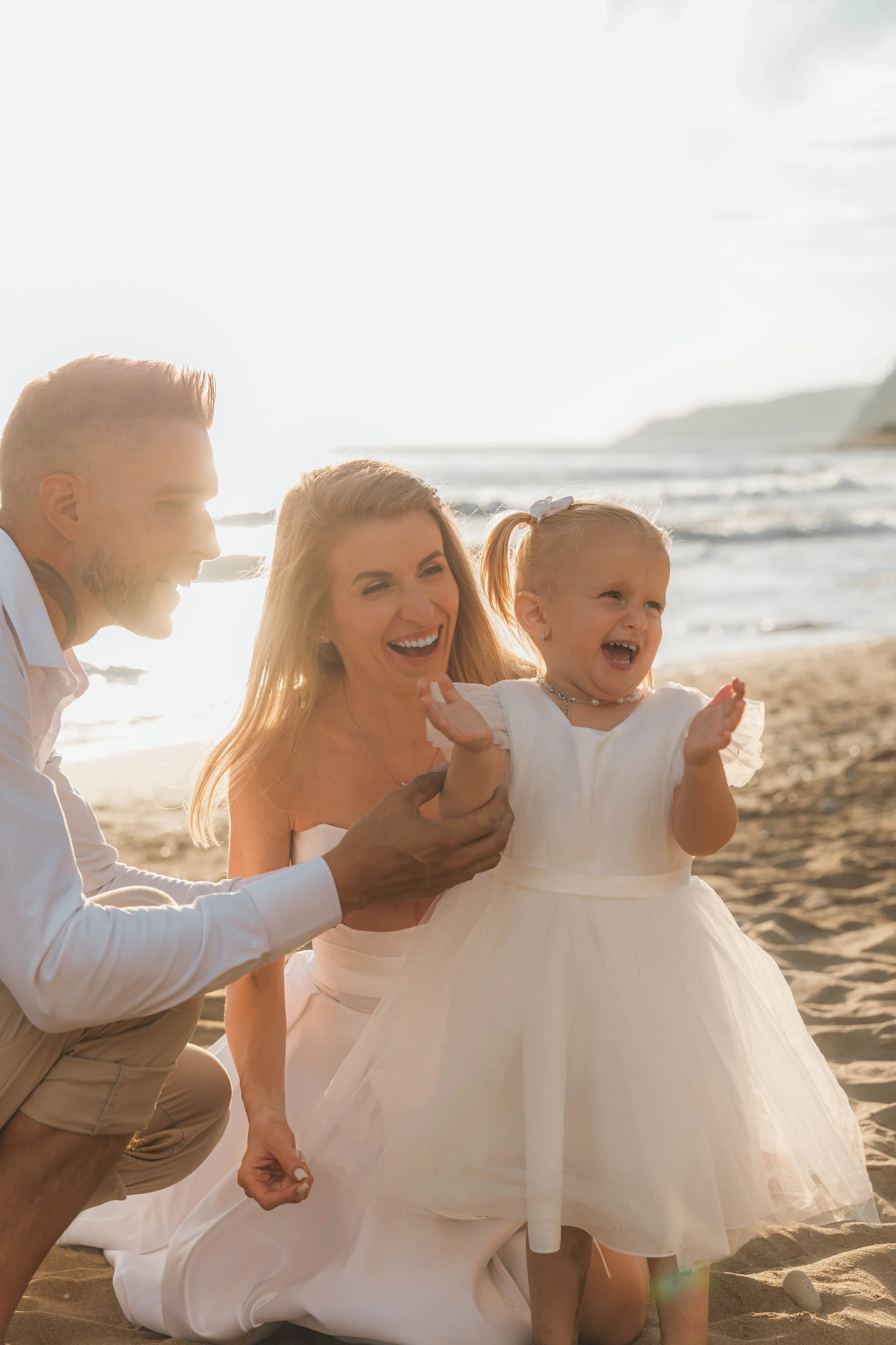 A family enjoying a joyful moment on the beach, with a smiling woman, young girl in a white dress, and man in casual attire, all laughing and playing together by the shoreline at sunset.