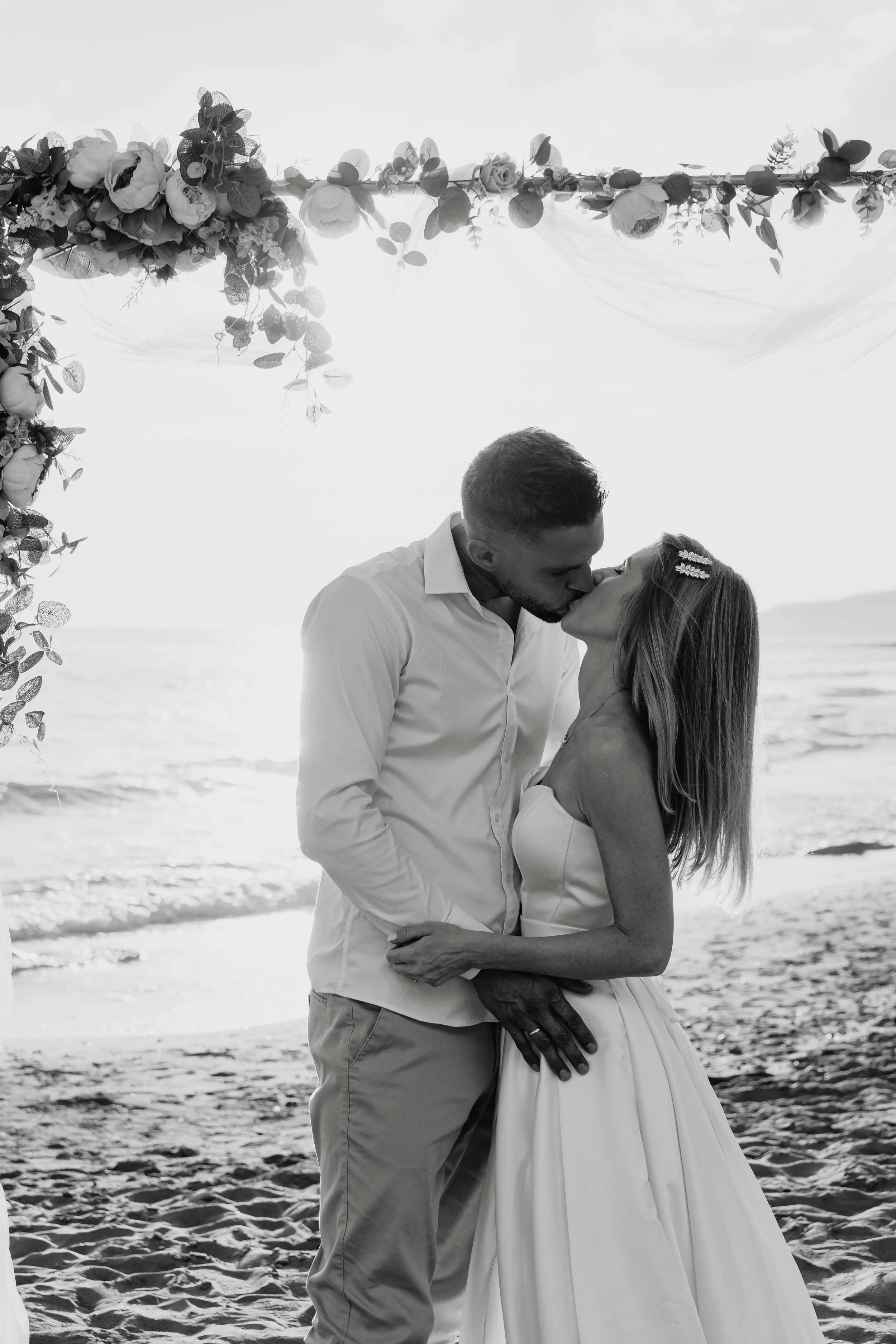 A black and white photo of a couple kissing on the beach, with an arch decorated with flowers and foliage overhead, sunset in the background.