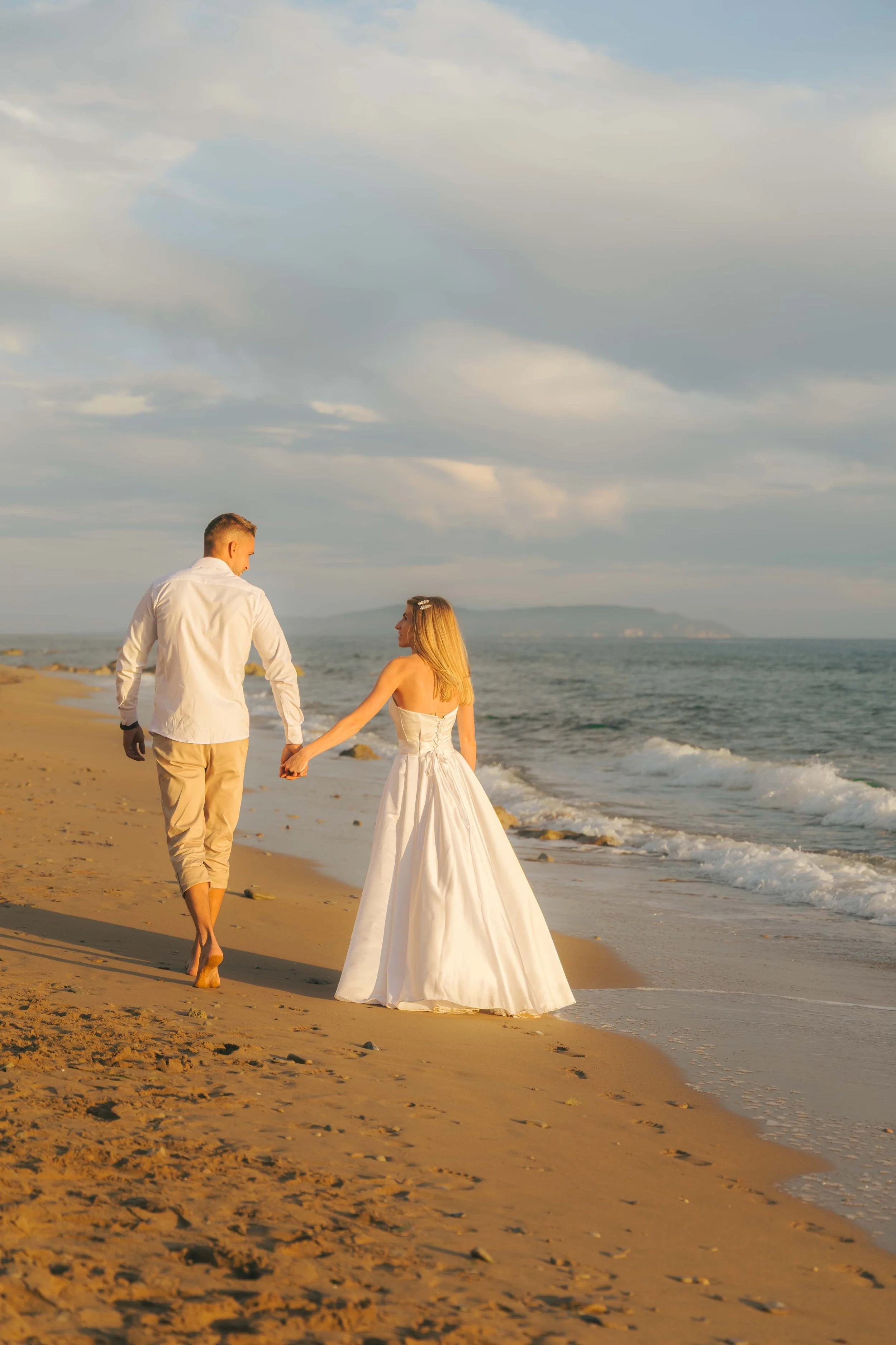A couple dressed in wedding attire holding hands walking along the beach at sunset.