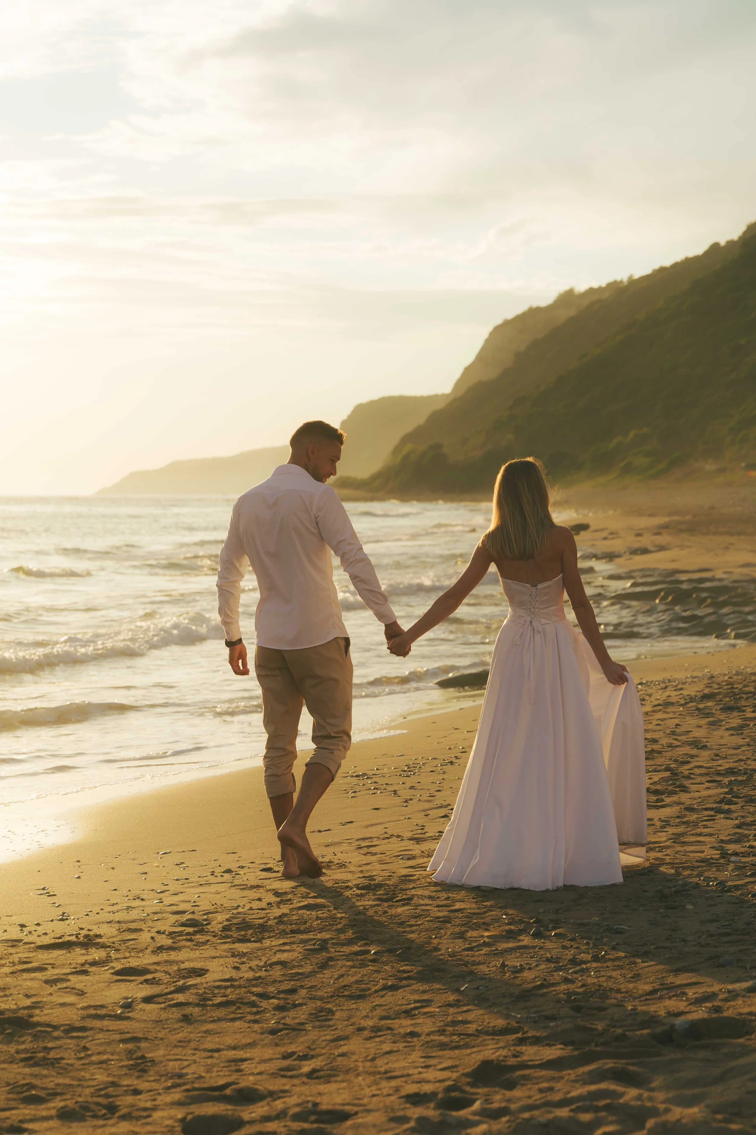 A man and woman walking hand in hand along a beach at sunset, with hills in the background and partially cloudy sky.