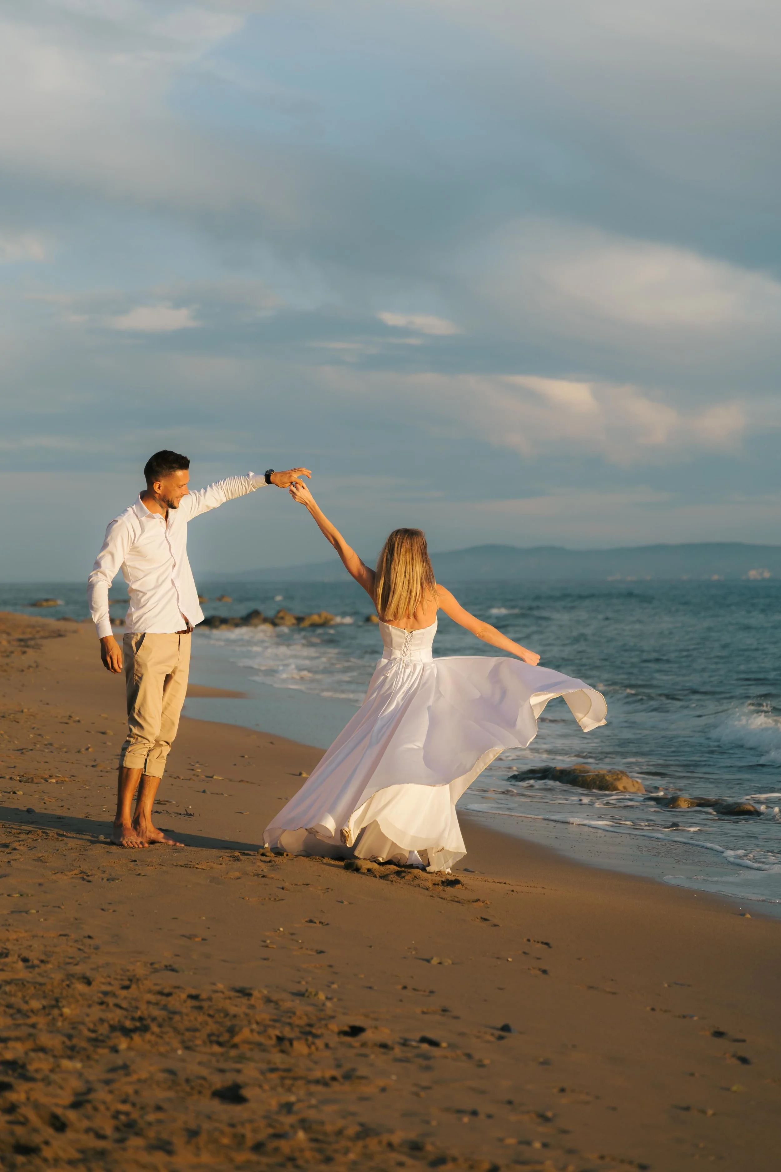 A couple dancing on the beach at sunset, with one woman wearing a white wedding dress and the man dressed in a white shirt and tan pants.