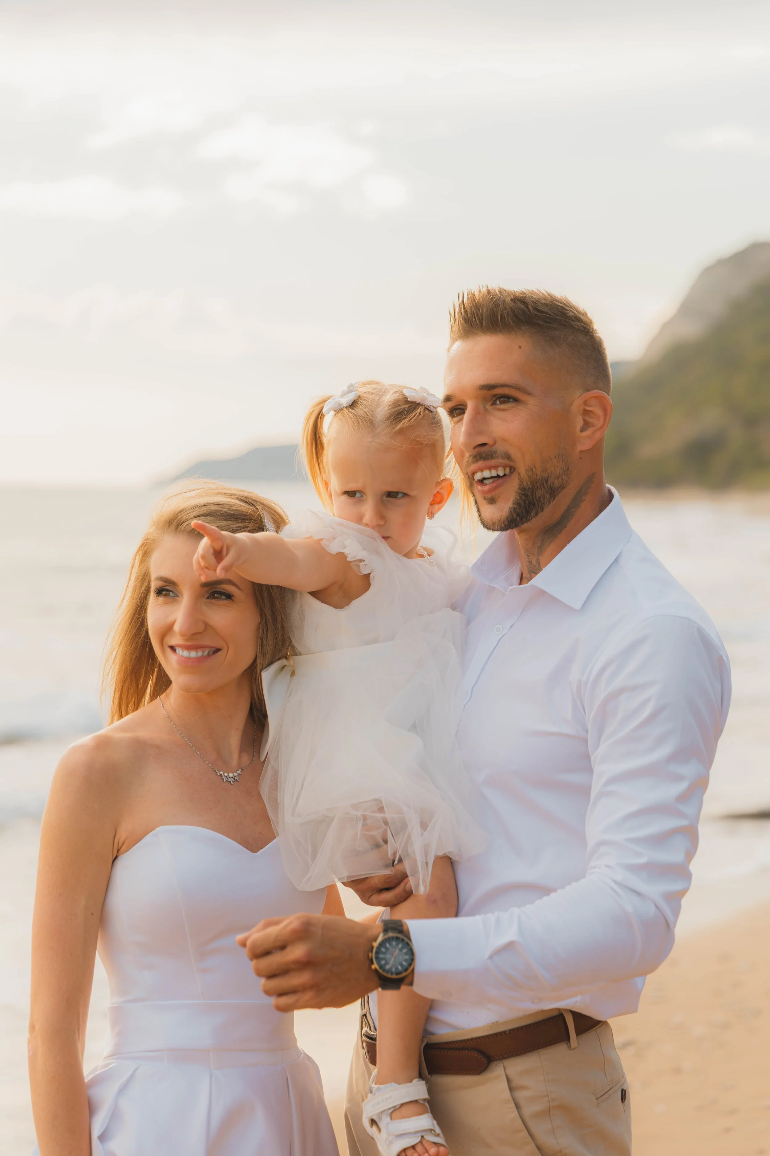 A family of three on a beach during sunset. The woman is wearing a white strapless dress, the man is in a white shirt and beige pants, holding a young girl in a white dress.