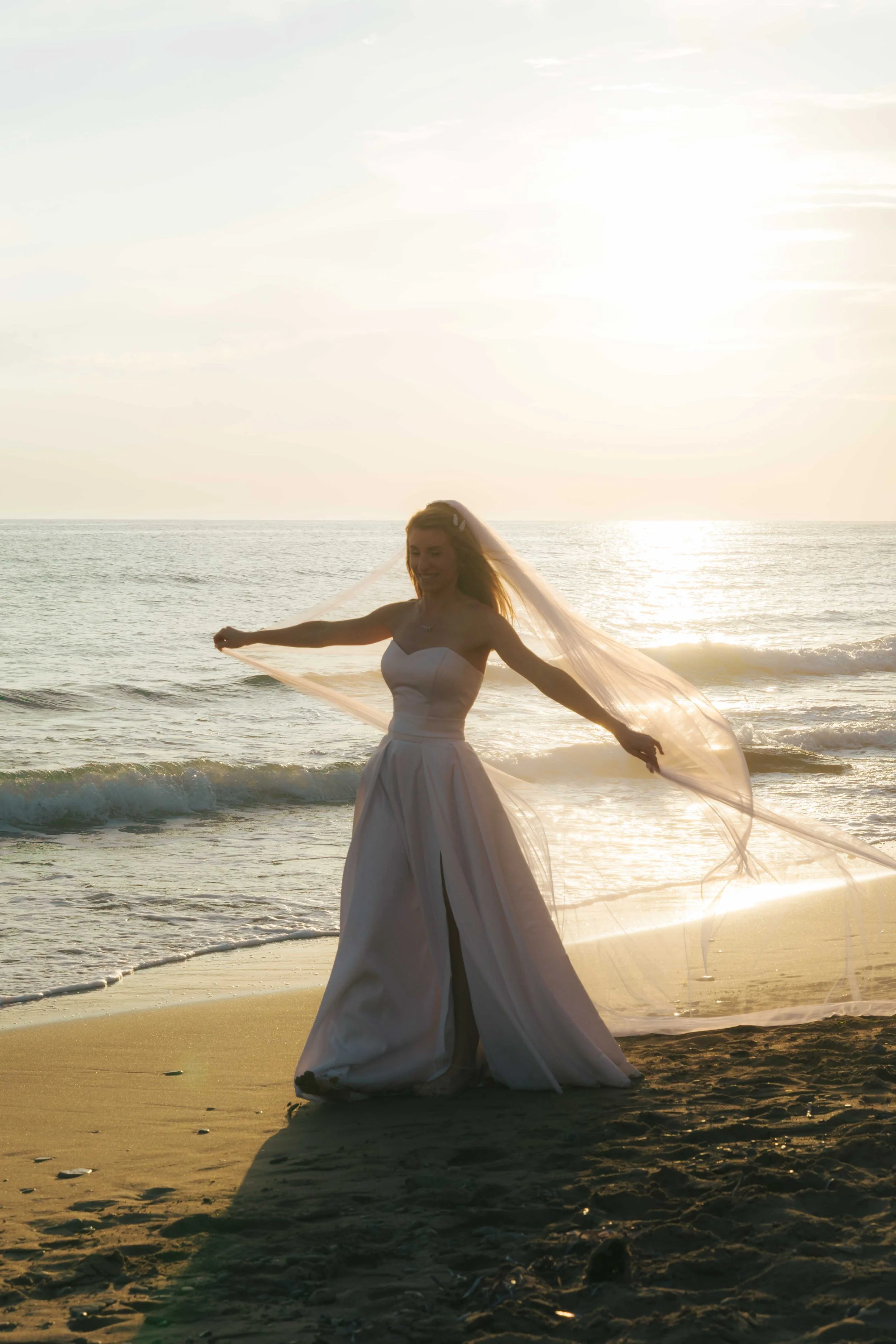 A bride in a white wedding dress holds a long veil on a sandy beach at sunset, with gentle waves and the ocean in the background.