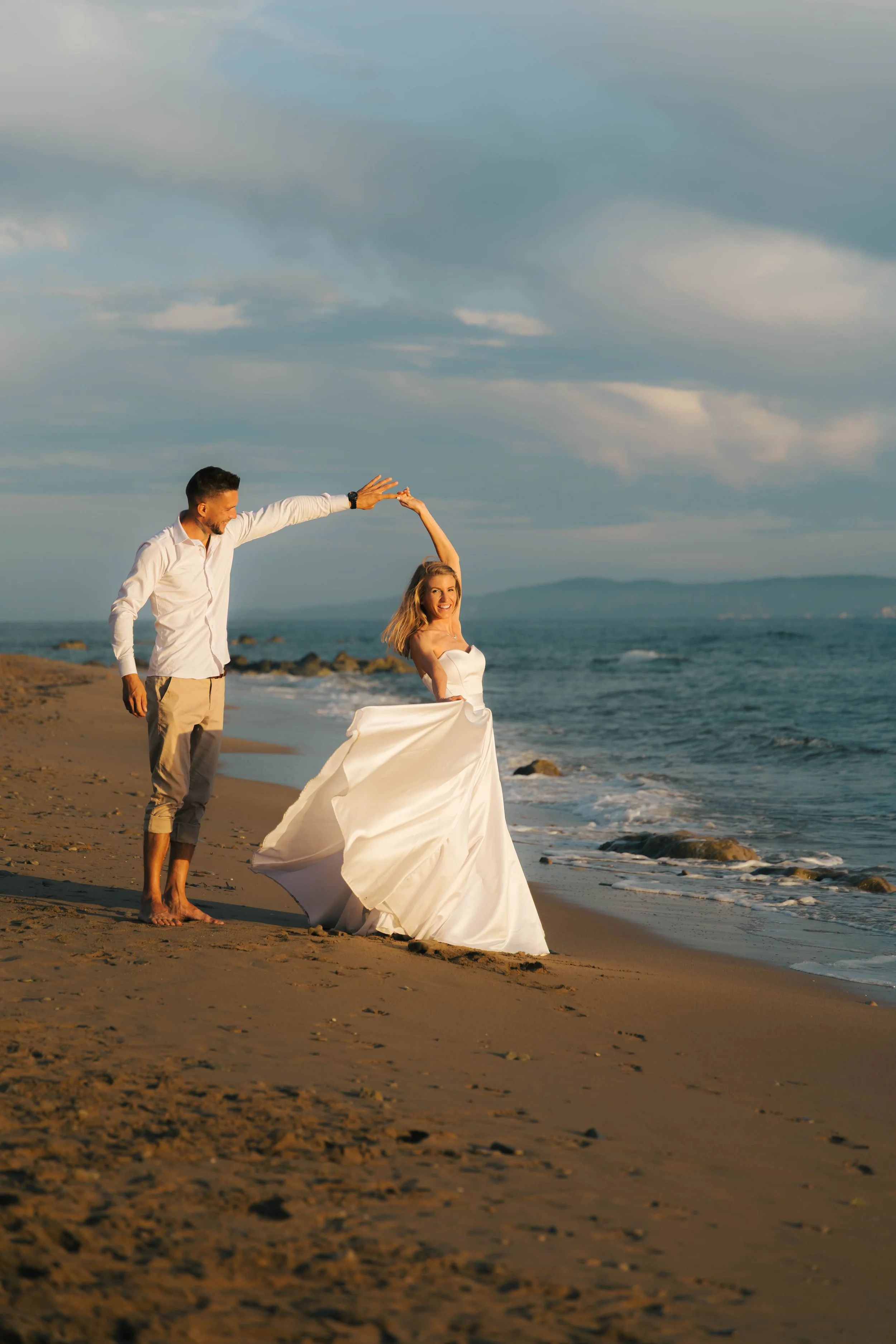 A happy couple on a beach, with the man holding the woman's hand as she spins in her wedding dress. The sky is partly cloudy, and the ocean is in the background.