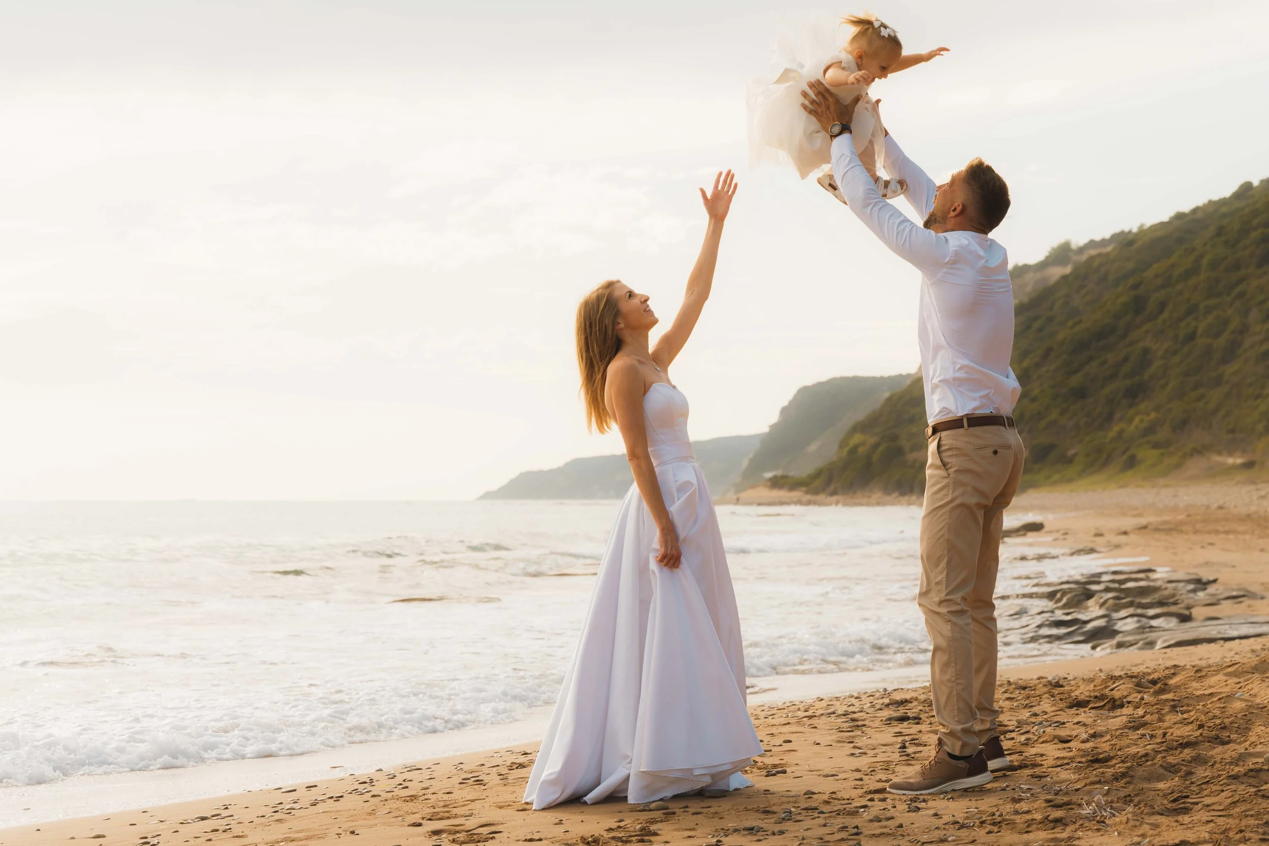 A couple, possibly newlyweds, on a beach with a young girl, likely their daughter. The woman, in a white dress, reaches out as the man lifts a little girl in a white dress, suggesting a family moment at sunset.