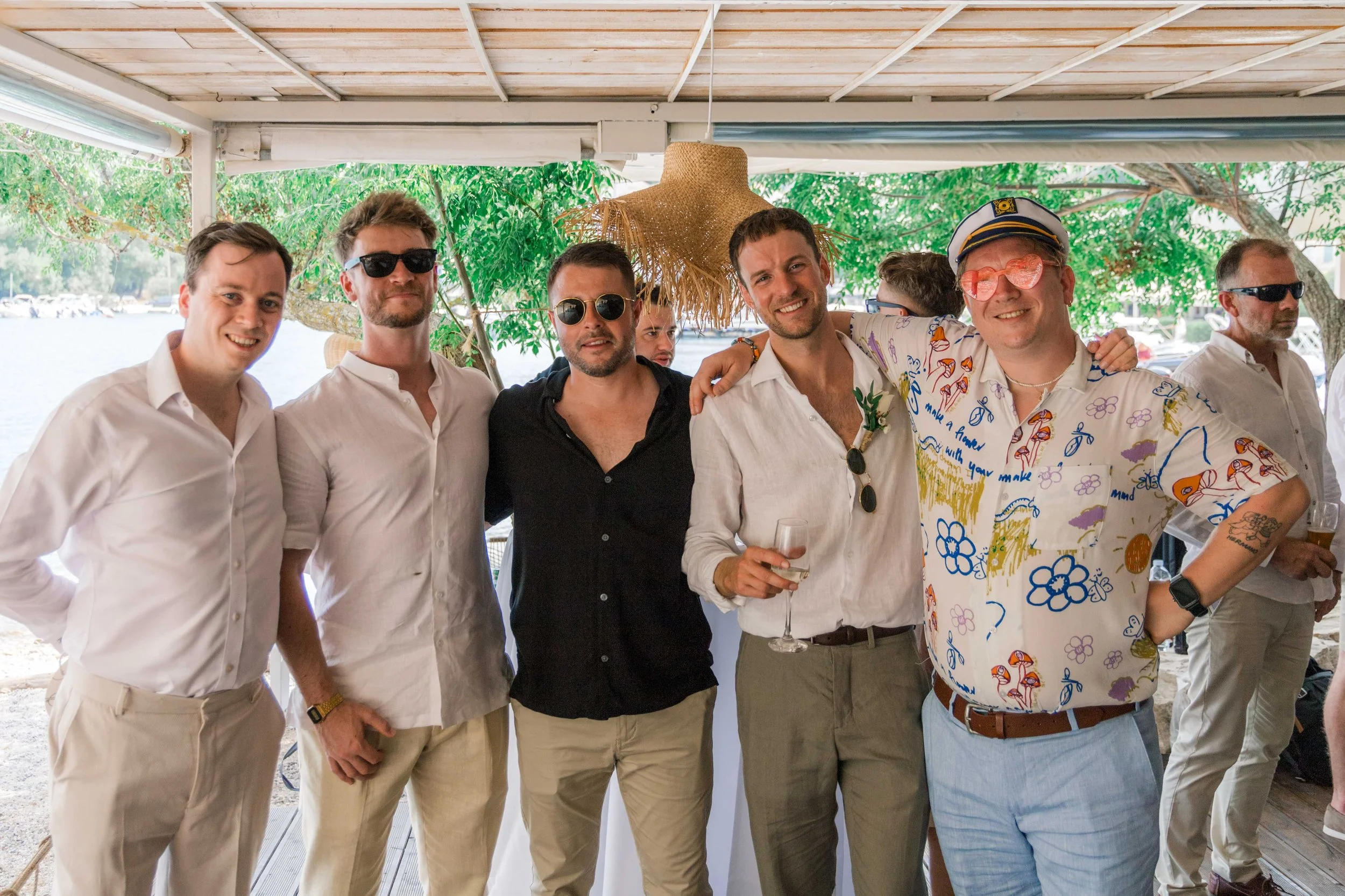 Group of men at a social gathering outdoors, some wearing sunglasses, with a decorative straw hat and tropical shirt, smiling and holding drinks.