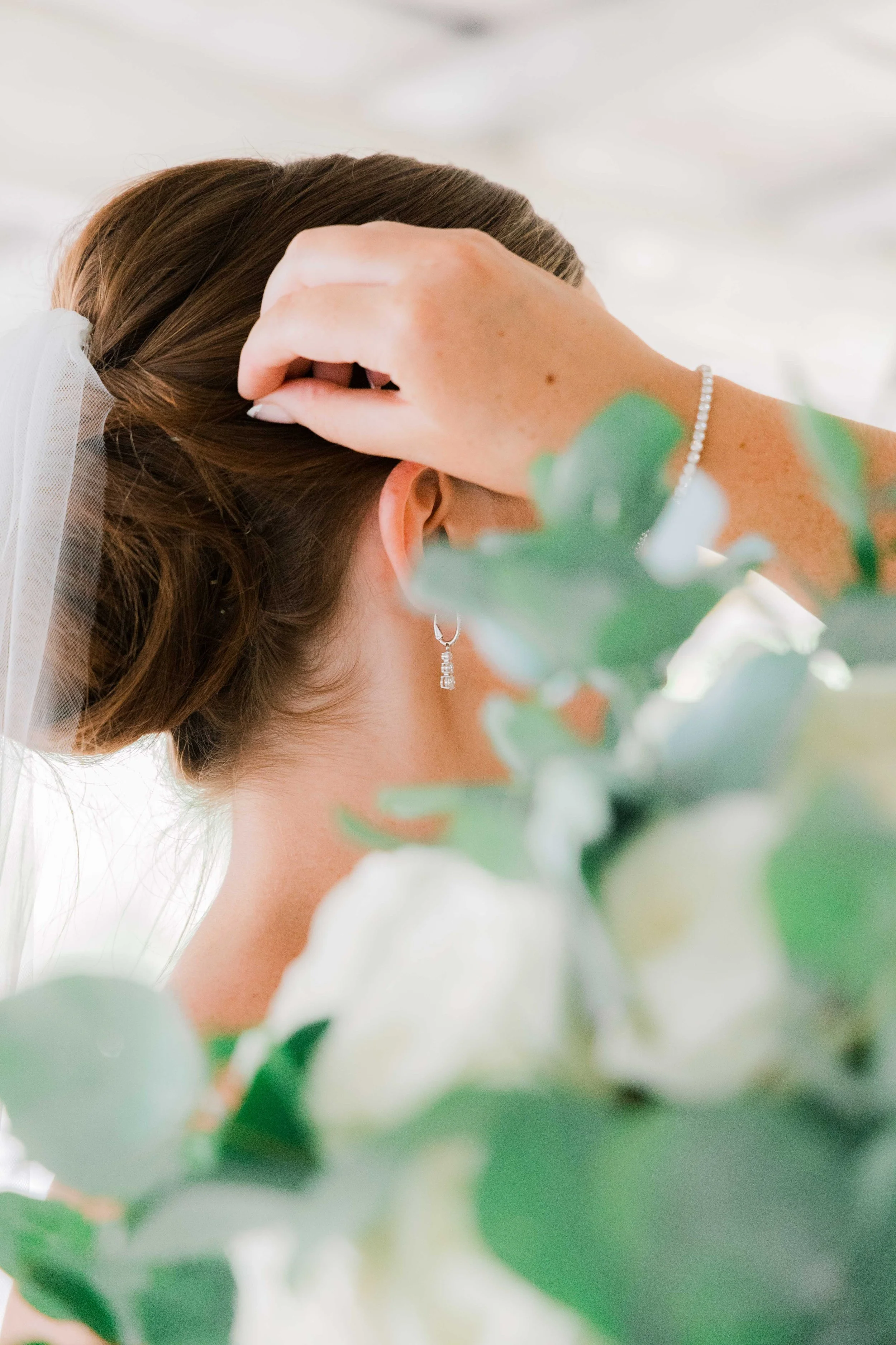 Close-up of a bride with a veil, earrings, and a bracelet, touching her hair with one hand, surrounded by flowers and greenery.