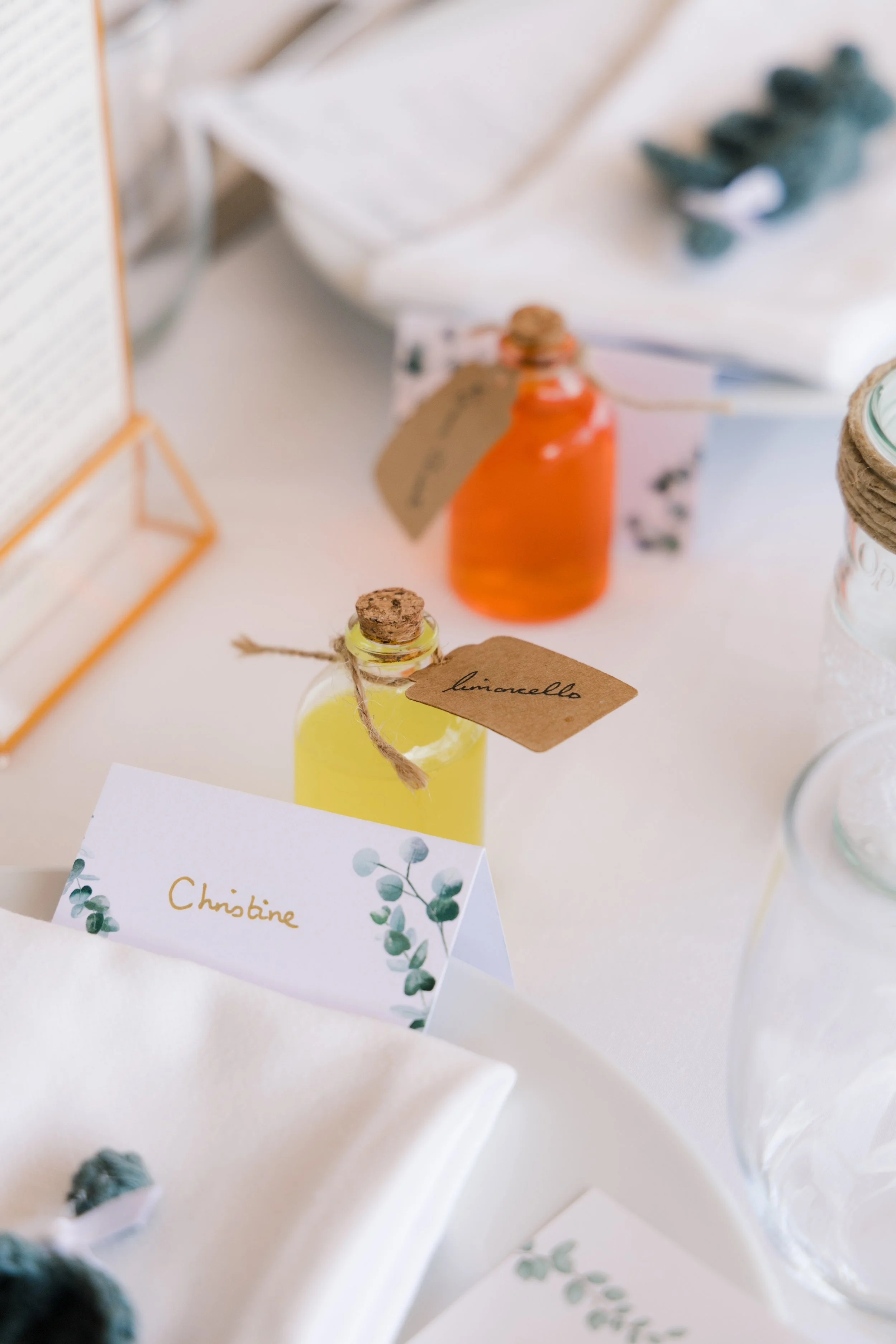 Table decor with small bottles of colorful liquids labeled 'limoncello' and 'amaretto,' and a place card with the name 'Christine' surrounded by greenery illustrations.
