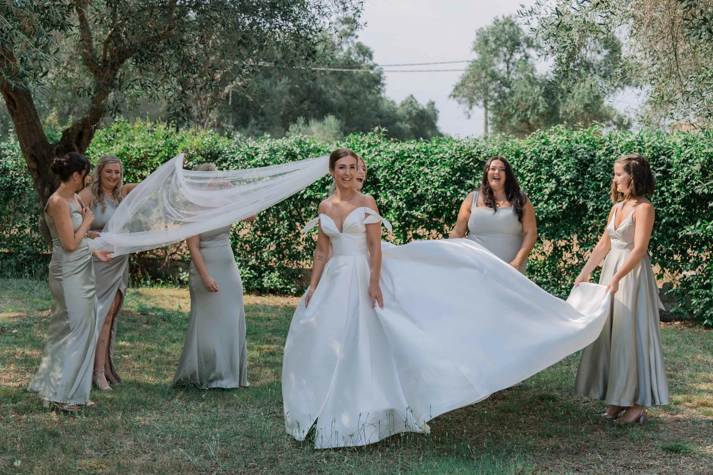 A bride in a white wedding gown surrounded by bridesmaids in matching silver dresses outdoors, with trees and bushes in the background.
