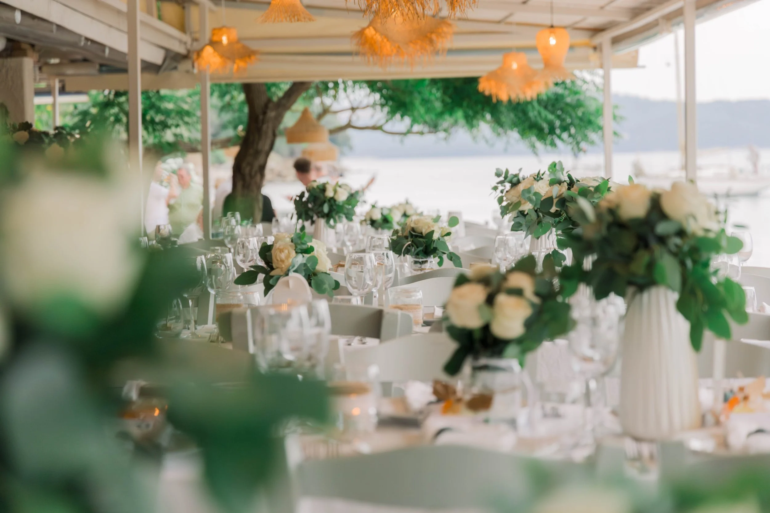 A decorated outdoor event space near water with white flowers, glassware, and table settings under a canopy with hanging lights.