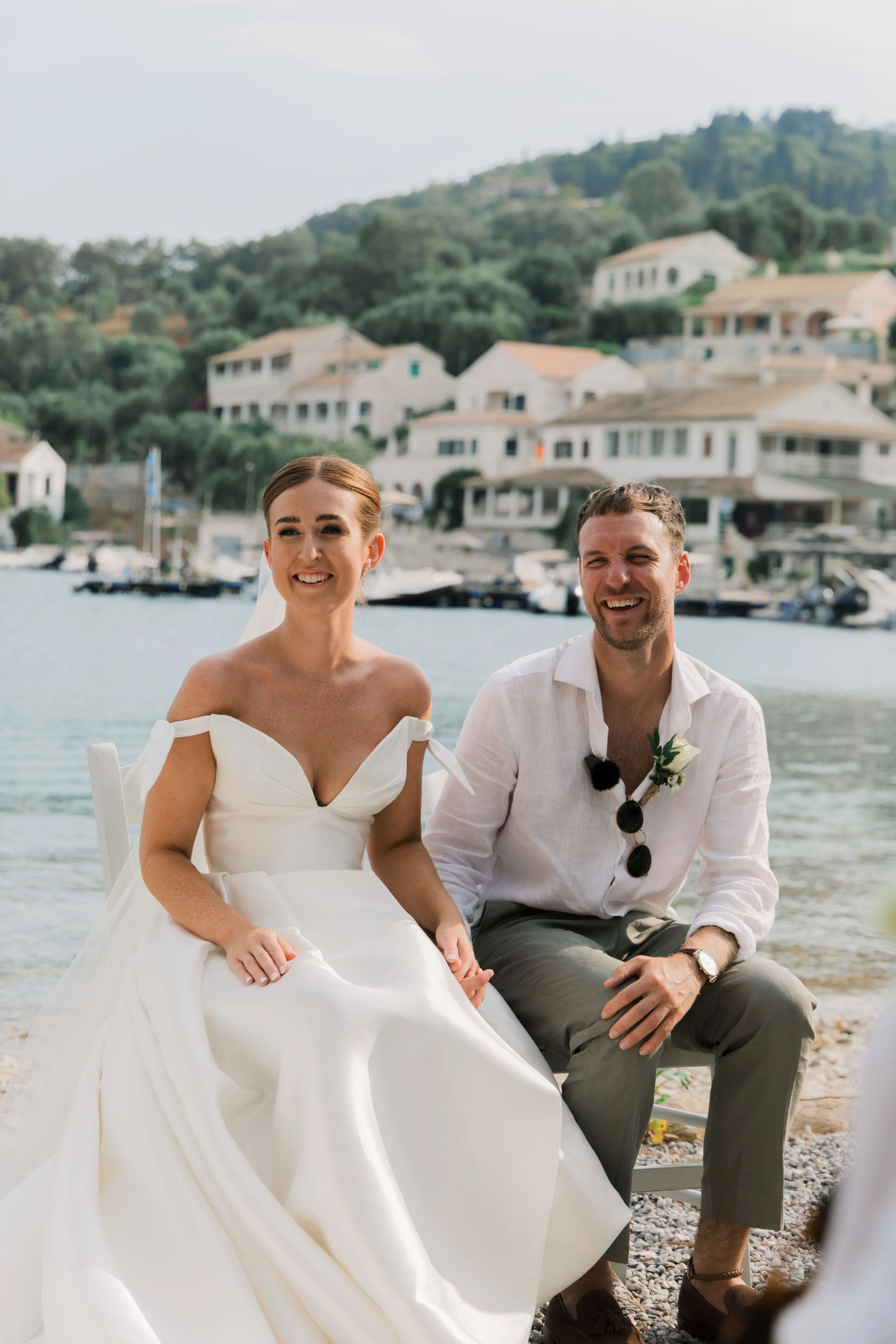 A bride and groom are sitting outdoors on a beach, smiling, with a waterfront background of houses and hills.