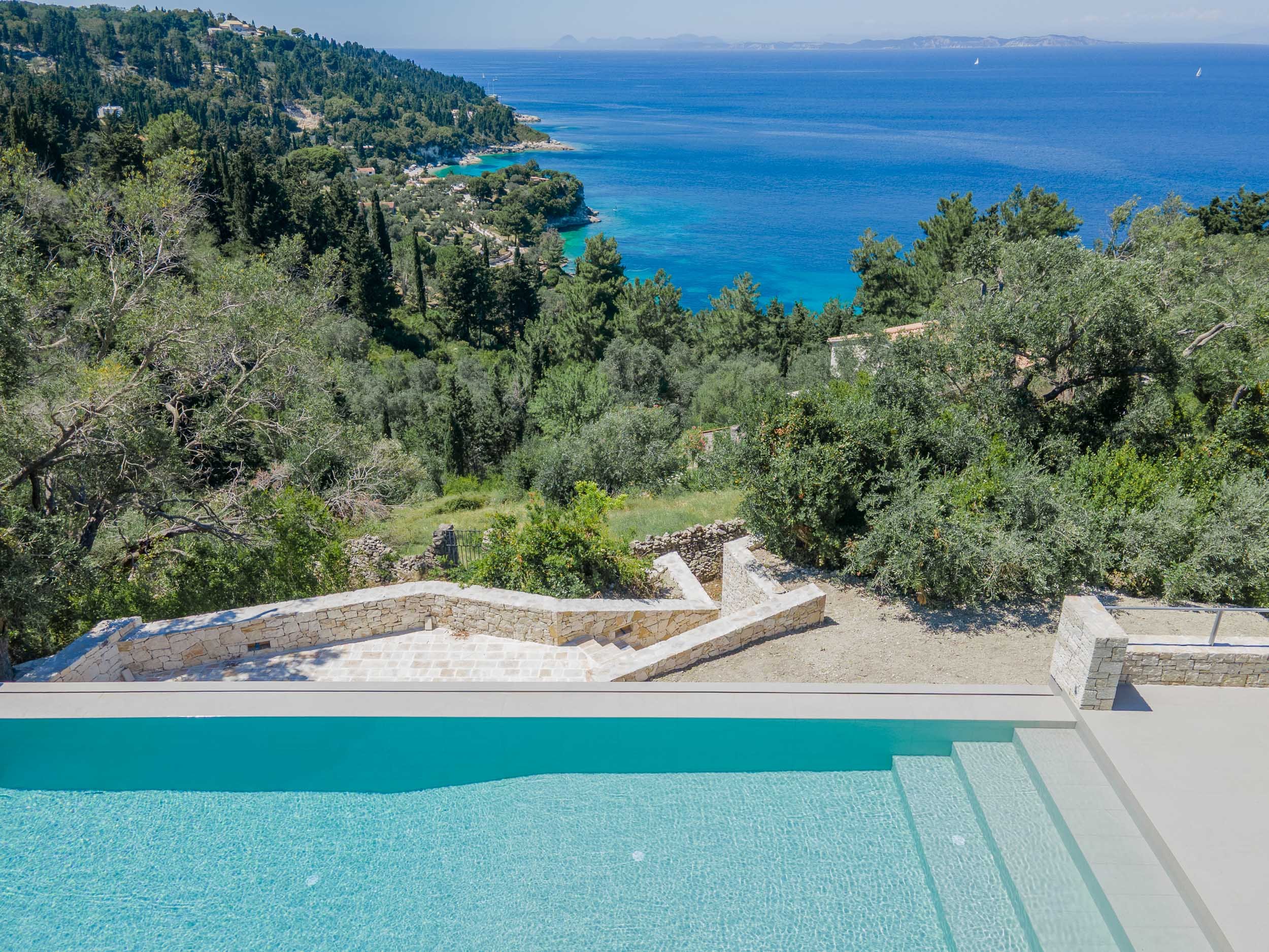 A swimming pool with a view of the ocean, surrounded by lush green trees and overlooking a hillside with houses and a distant coastline.