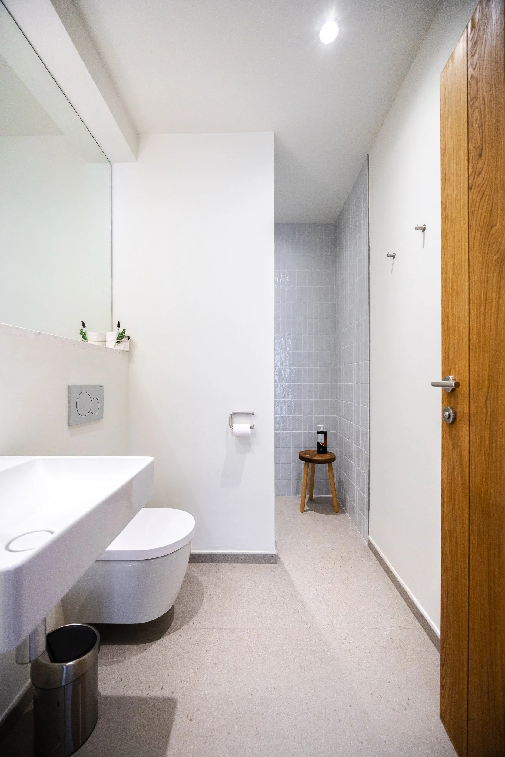 A clean, modern bathroom with a white ceramic toilet, a small sink, a mirror, three small potted plants, a metal trash can, a wooden stool with a bottle on top, hooks on the wall, and a separate shower area with white tiles, a round seat, and a bottl