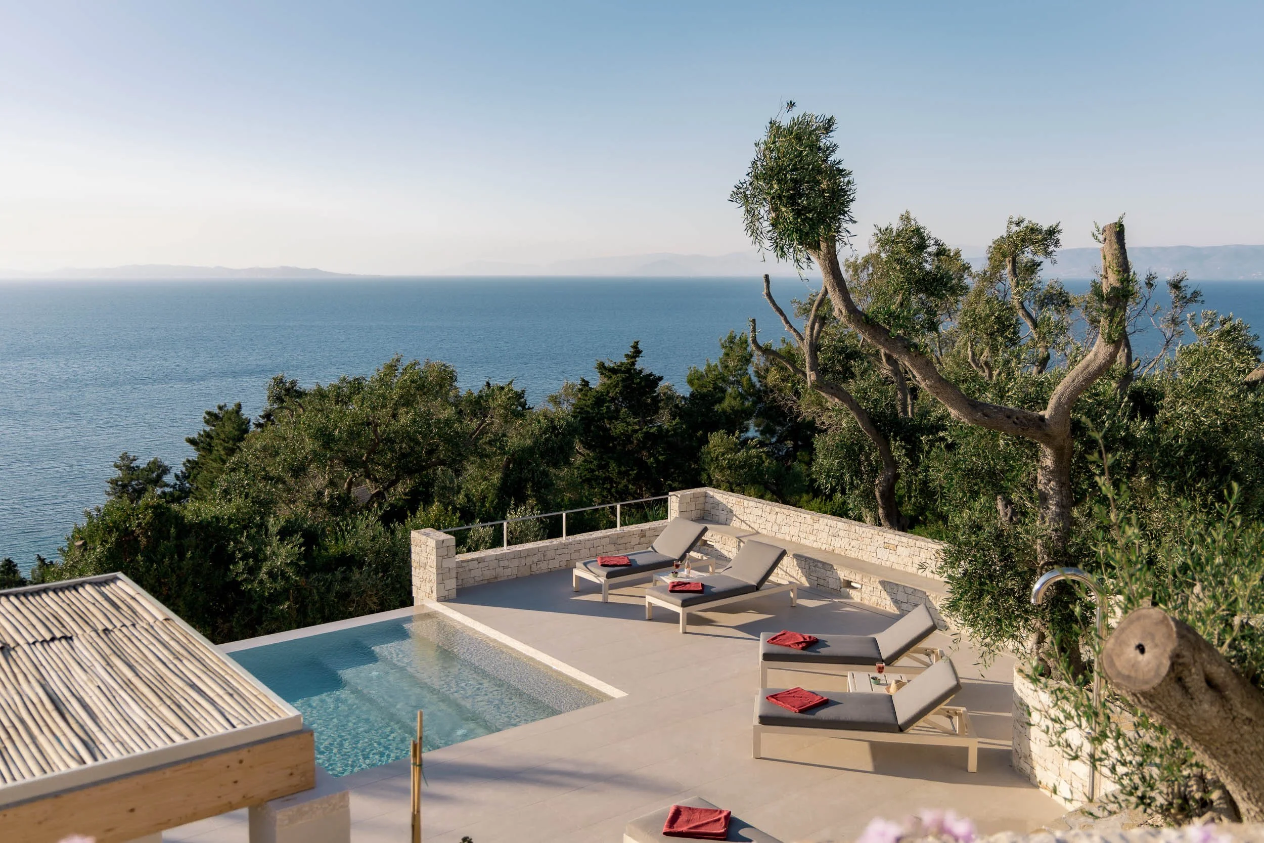 Luxury outdoor pool area with four lounge chairs with red towels overlooking the ocean, surrounded by trees and a stone wall.