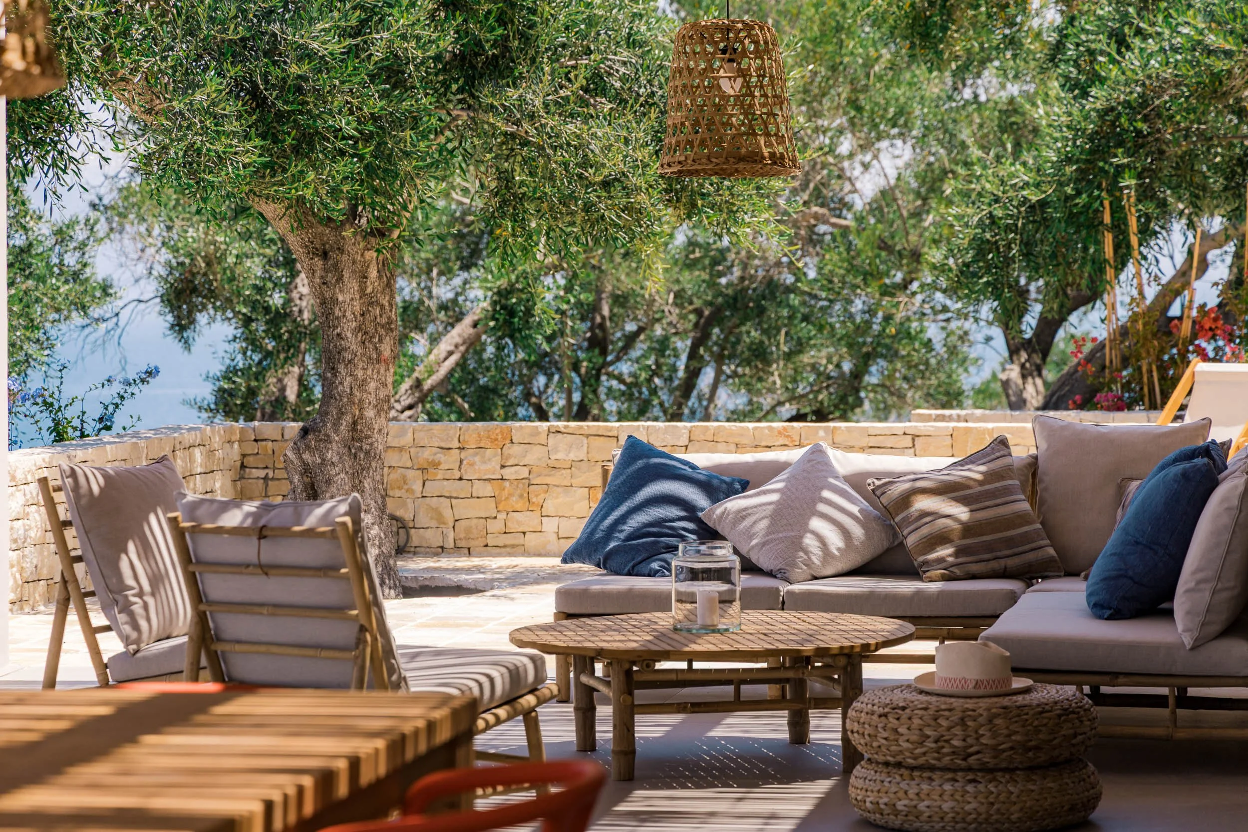 Outdoor patio with a stone wall, cushioned seating with pillows, a wooden coffee table with a candle, a tree with green foliage, and a hanging wicker lamp.