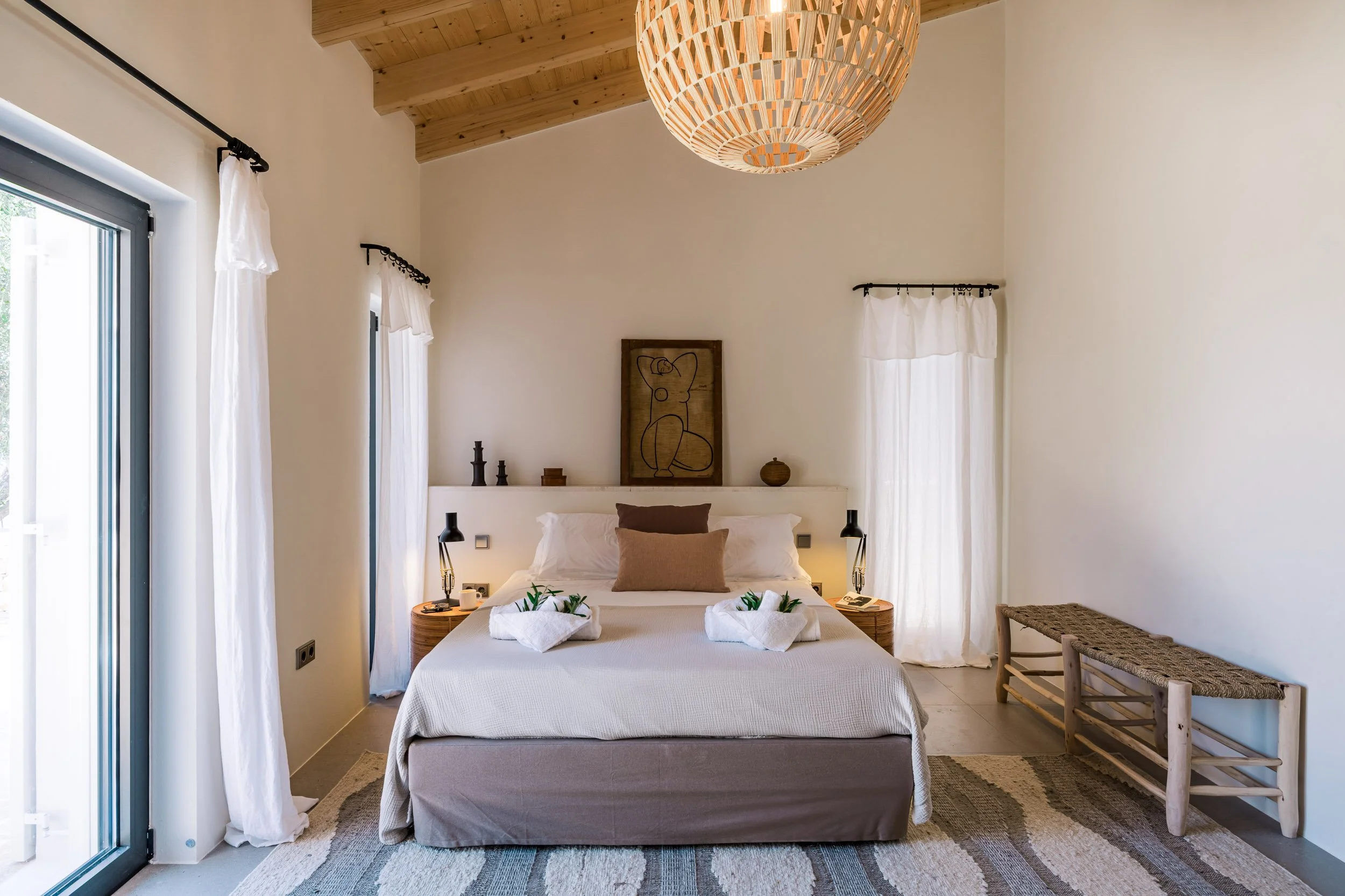 A bedroom with a neutral color scheme, featuring a bed with pillows and towels with greenery, two nightstands with lamps, a wooden bench, and natural light coming through white curtains, with a framed abstract drawing above the headboard.