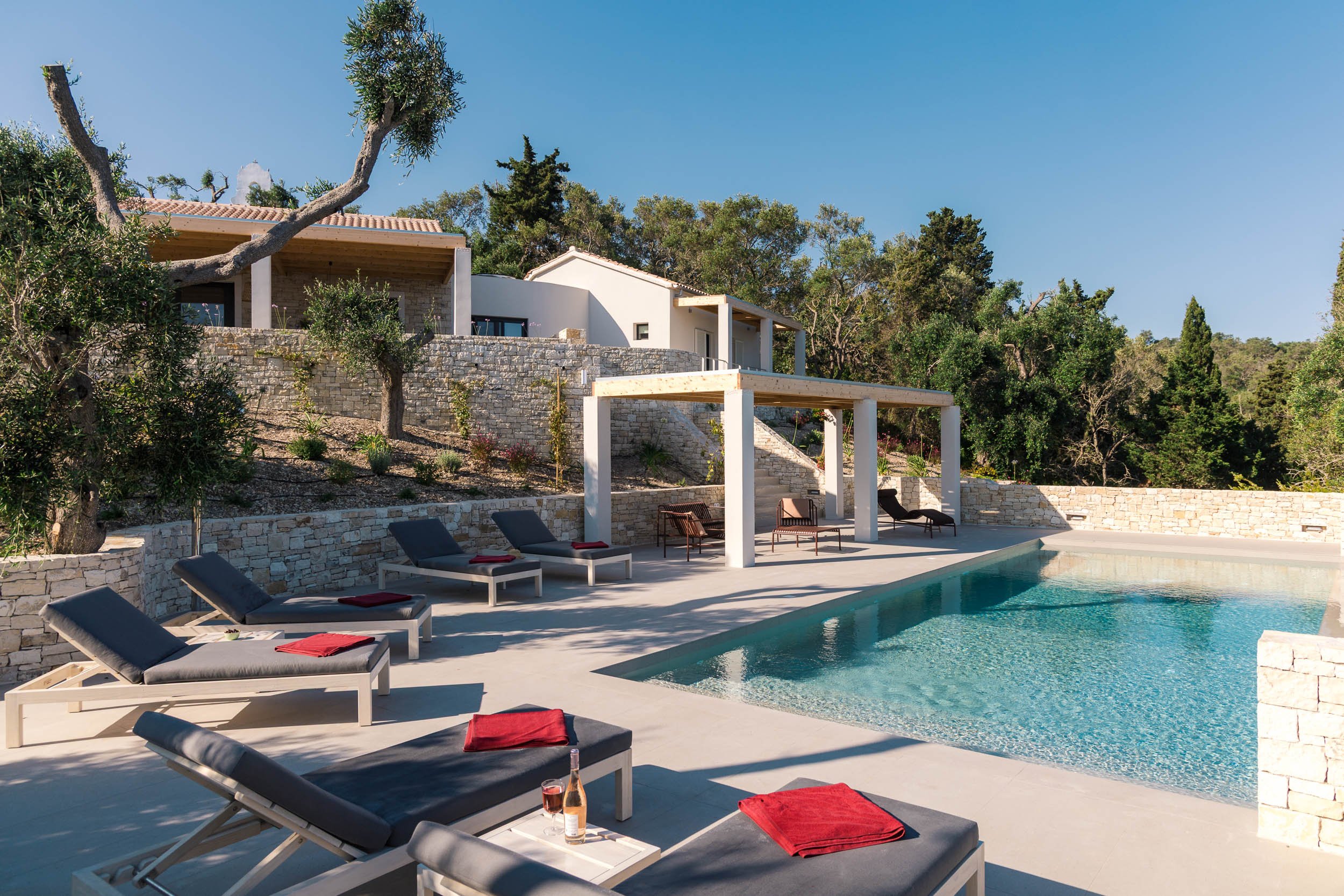 Luxury modern house with outdoor pool, lounge chairs with towels, and scenic green trees in the background under clear blue sky.