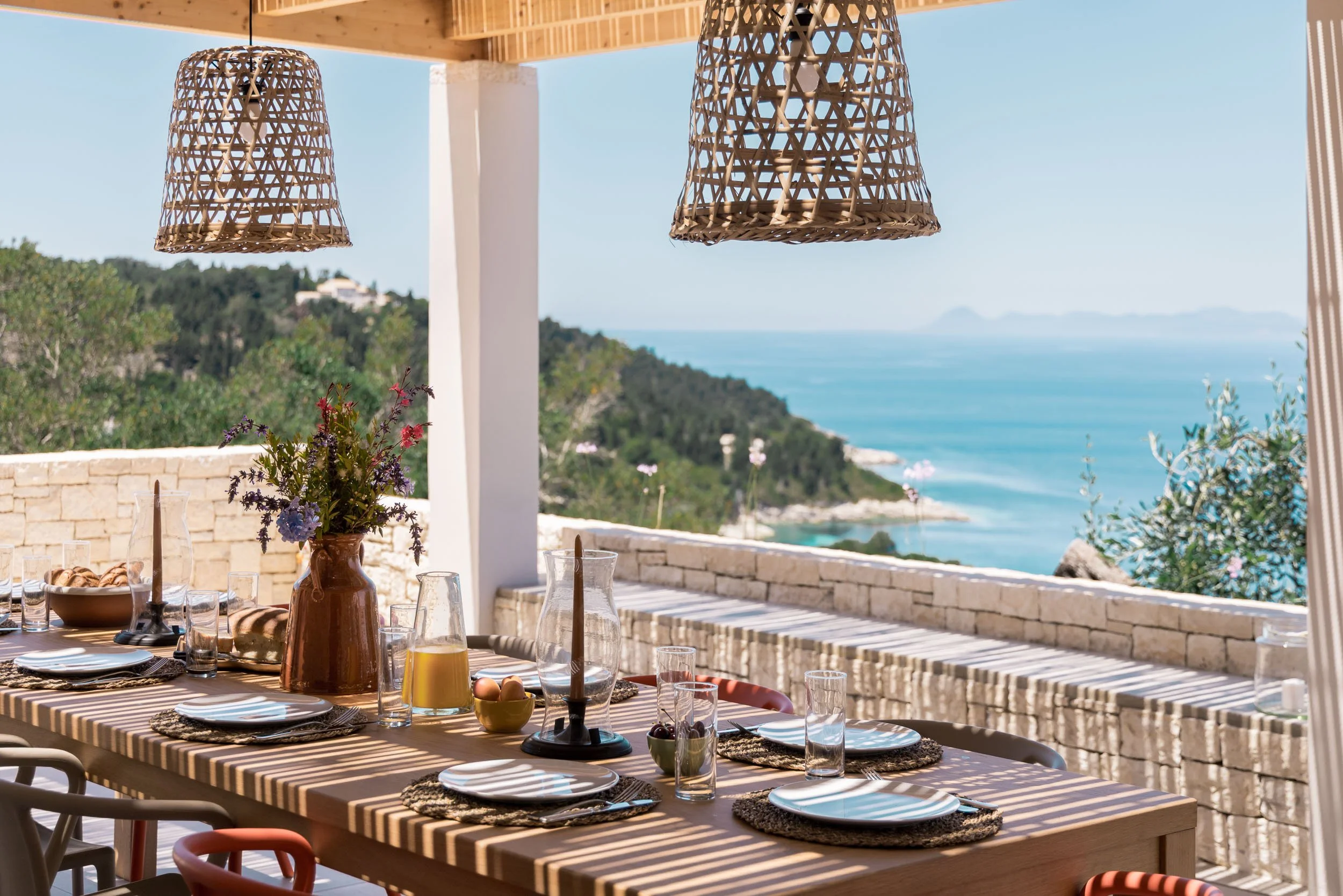 Outdoor dining table set for a meal, with plates, glasses, a vase with flowers, and candles, overlooking a scenic ocean view with hills and islands in the distance, under woven pendant lights.