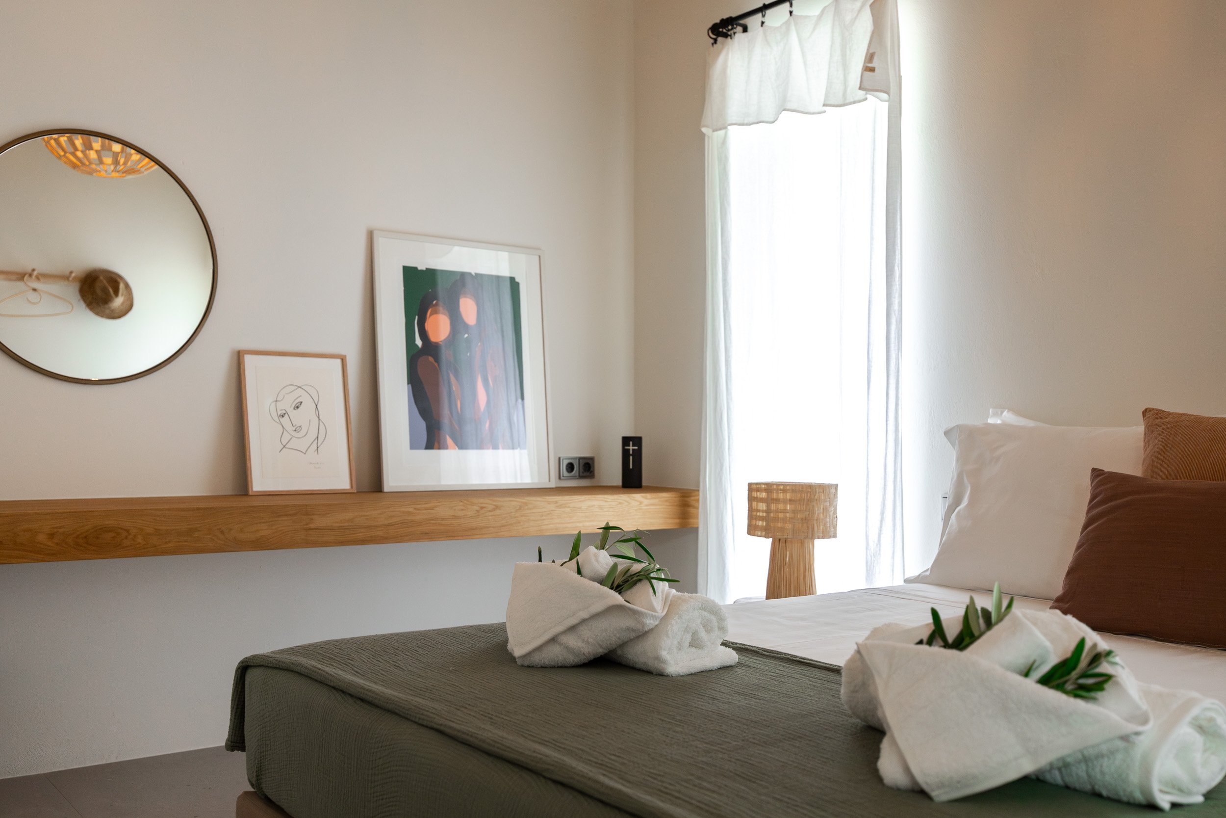 A neatly made bed with white linens, brown pillows, and towel art with greenery on top. A window with sheer white curtains allows natural light into the room. A wooden nightstand with a woven lamp sits beside the bed. There is a wooden ledge on the w