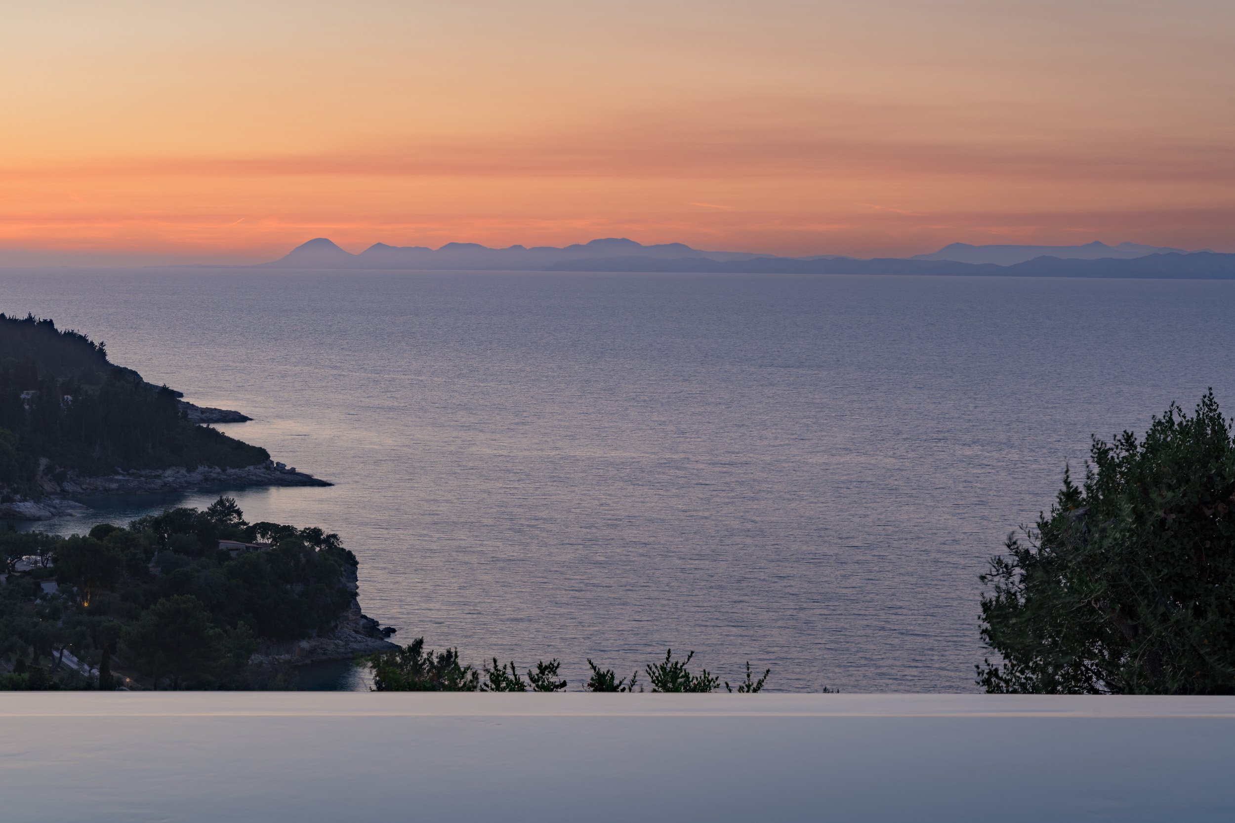 A sunset over a body of water with distant mountains in the background and some trees along the coastline in the foreground.