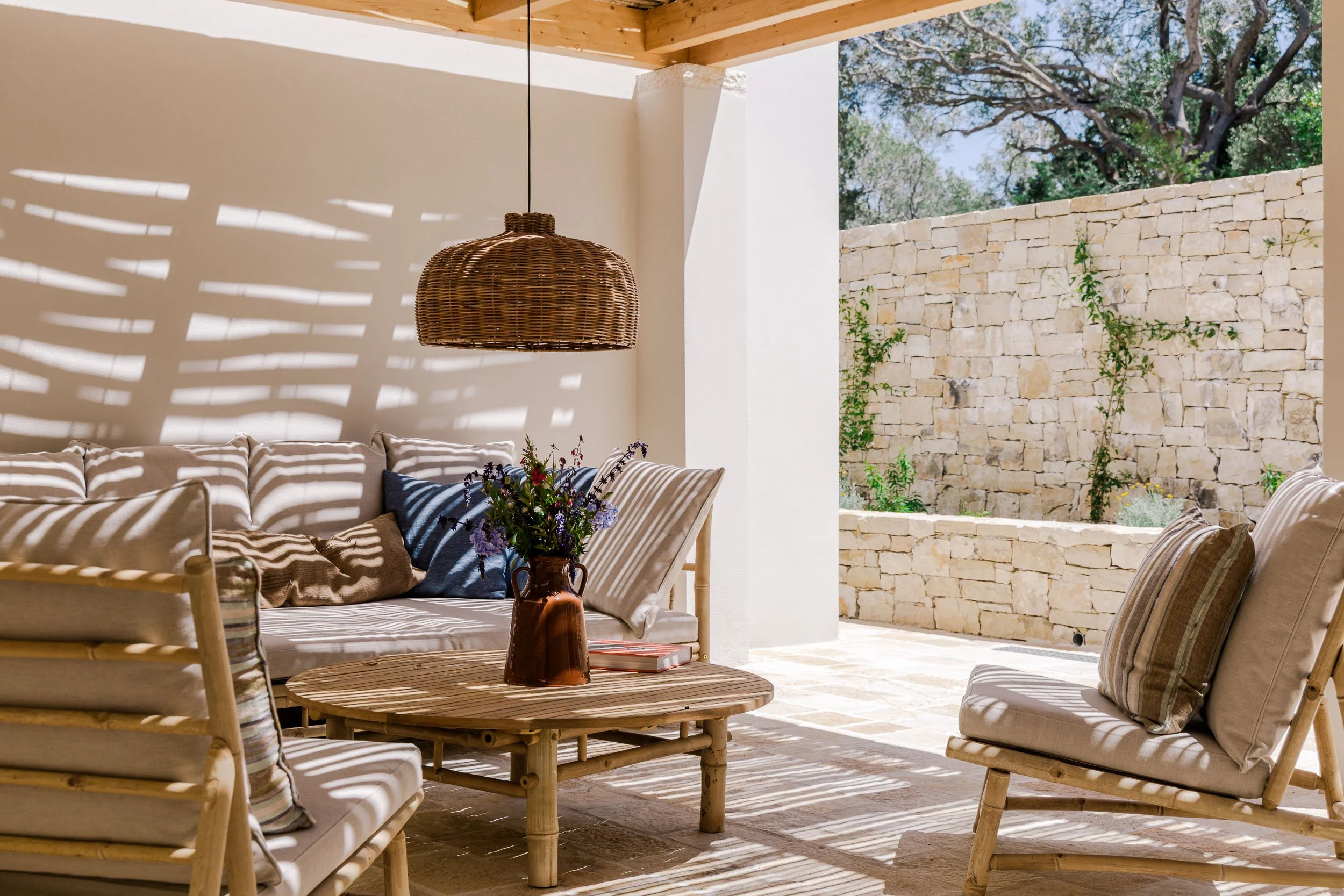 Outdoor patio with rattan furniture, cushions, a coffee table with a vase of flowers, and a view of a stone wall with plants, under a wooden ceiling with hanging rattan light fixture.