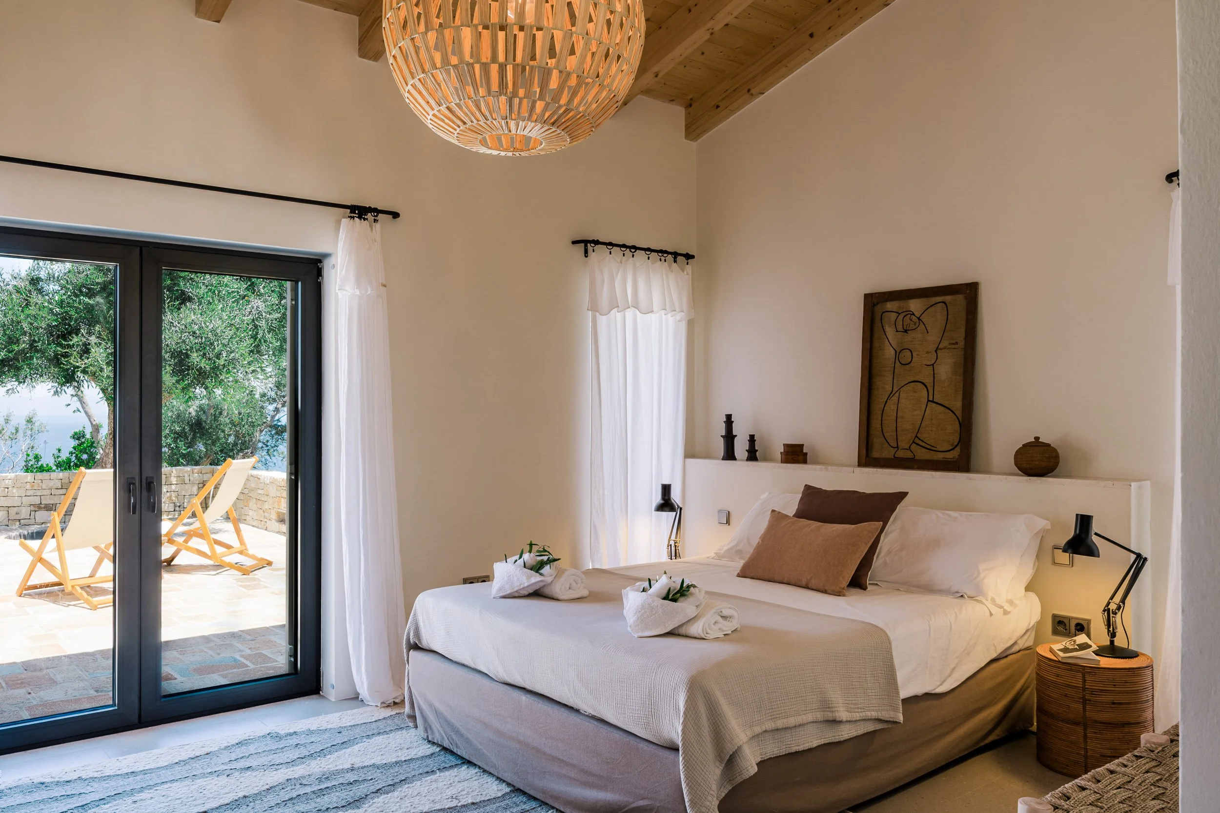 A bedroom with a large bed, brown pillows, and white linens, overlooking a balcony with outdoor chairs and lush greenery outside.
