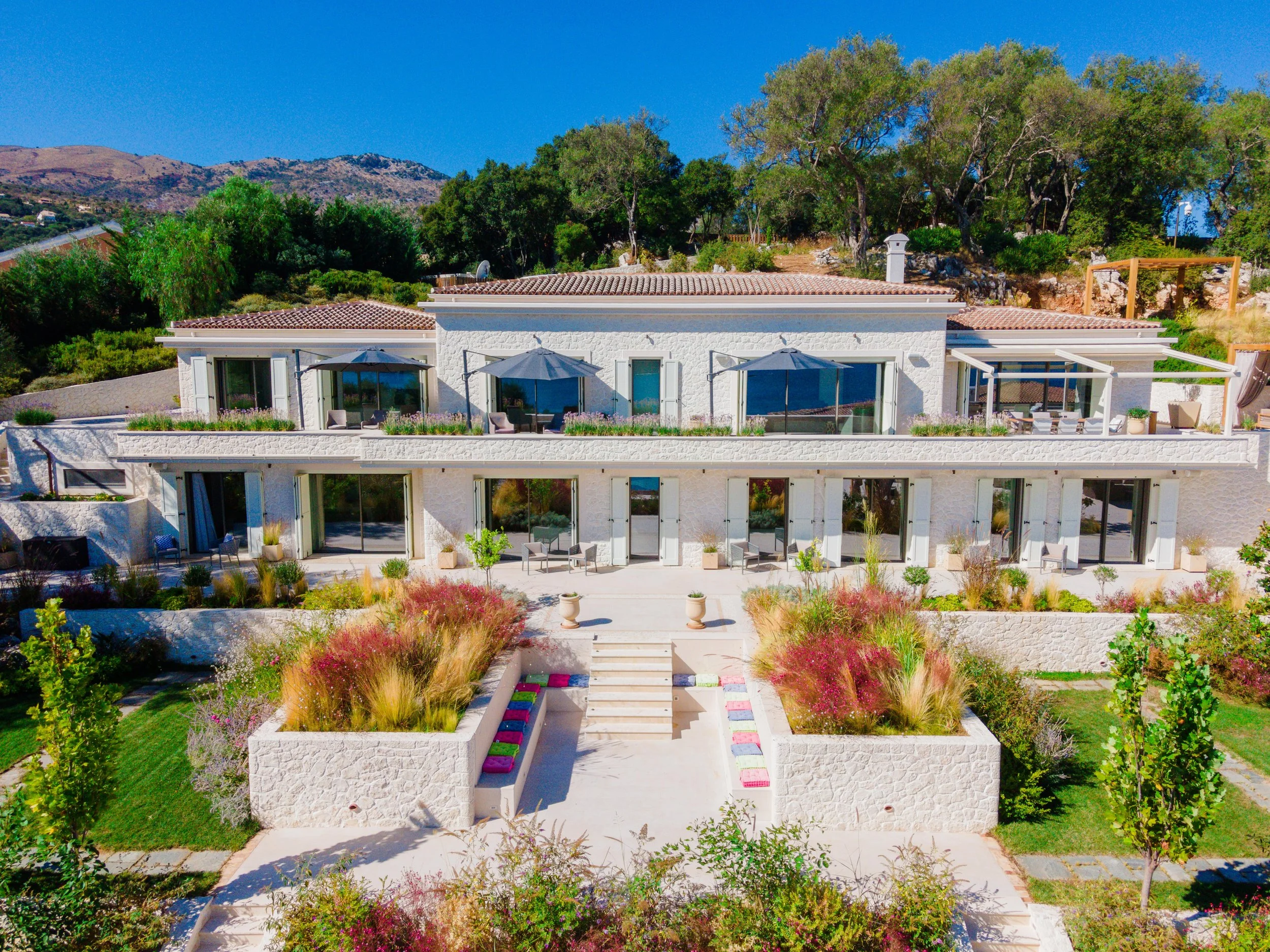 A modern, white multi-story house with large glass windows and sliding doors, surrounded by landscaped gardens with colorful plants and trees, set against a hilly background under a clear blue sky.