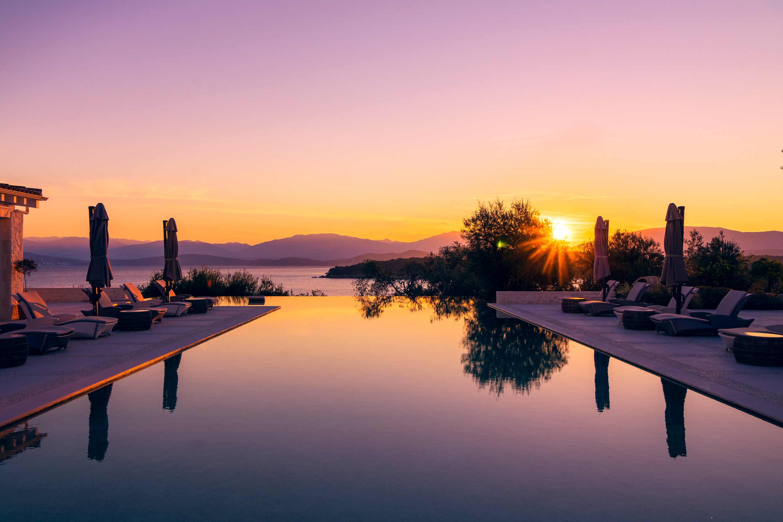 An infinity pool overlooking a lake and mountains during sunset with poolside lounge chairs and umbrellas.