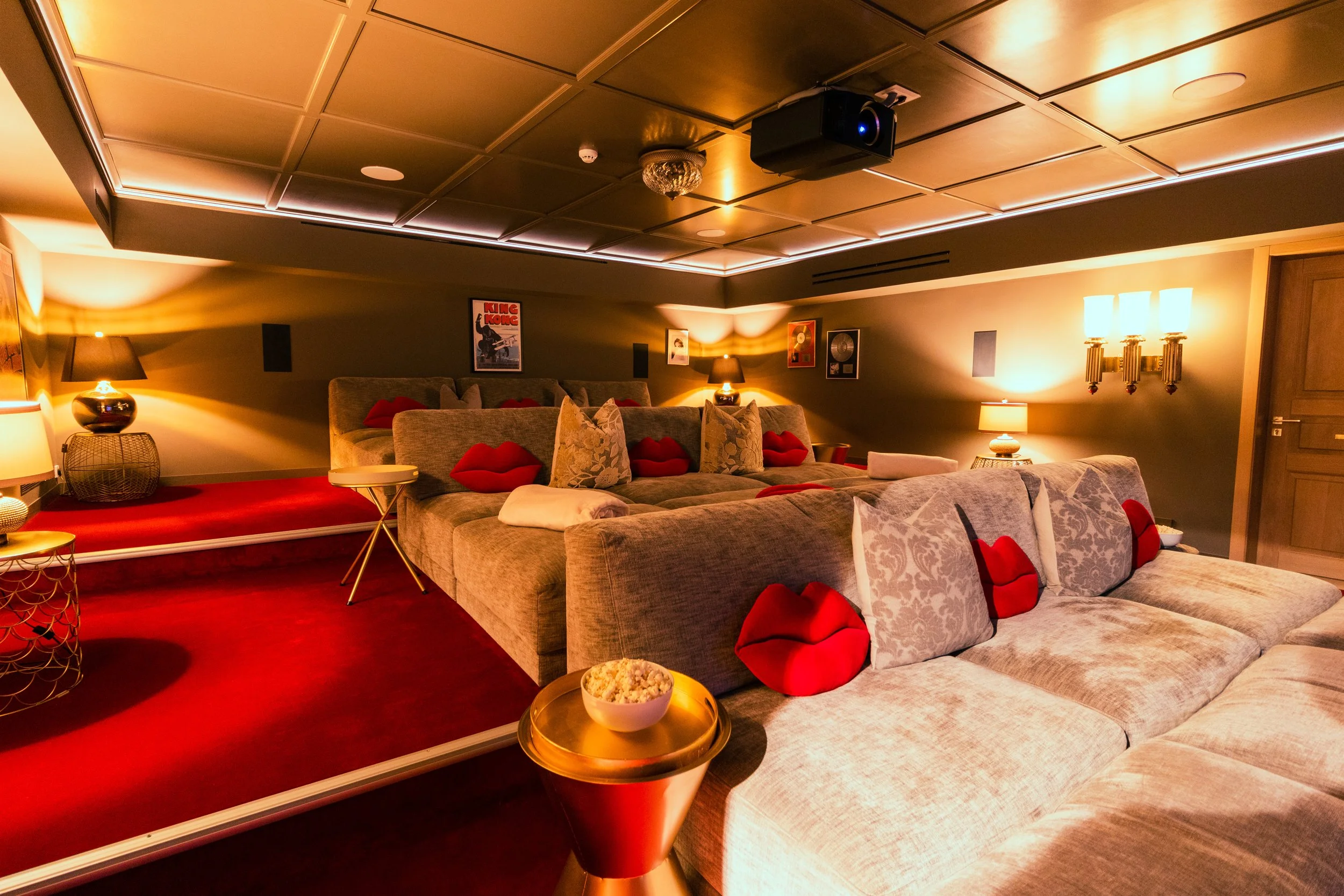 A cozy home theater room with gray couches decorated with red lips-shaped pillows and floral-patterned pillows, warm lighting, framed artwork on the walls, and a projector mounted on a paneled ceiling, with a red carpeted section for seating.