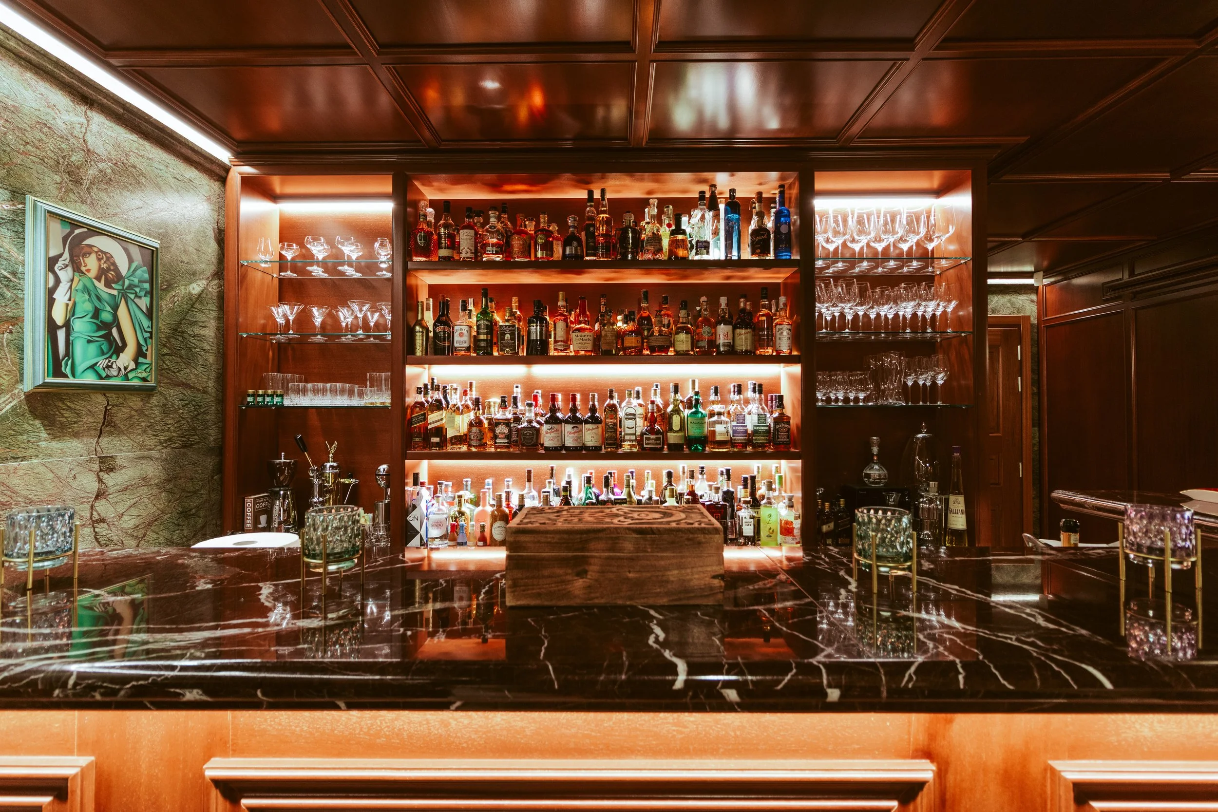 A bar with a dark marble counter and a wooden back shelf filled with various bottles of liquor, glassware, and illuminated with warm lighting.