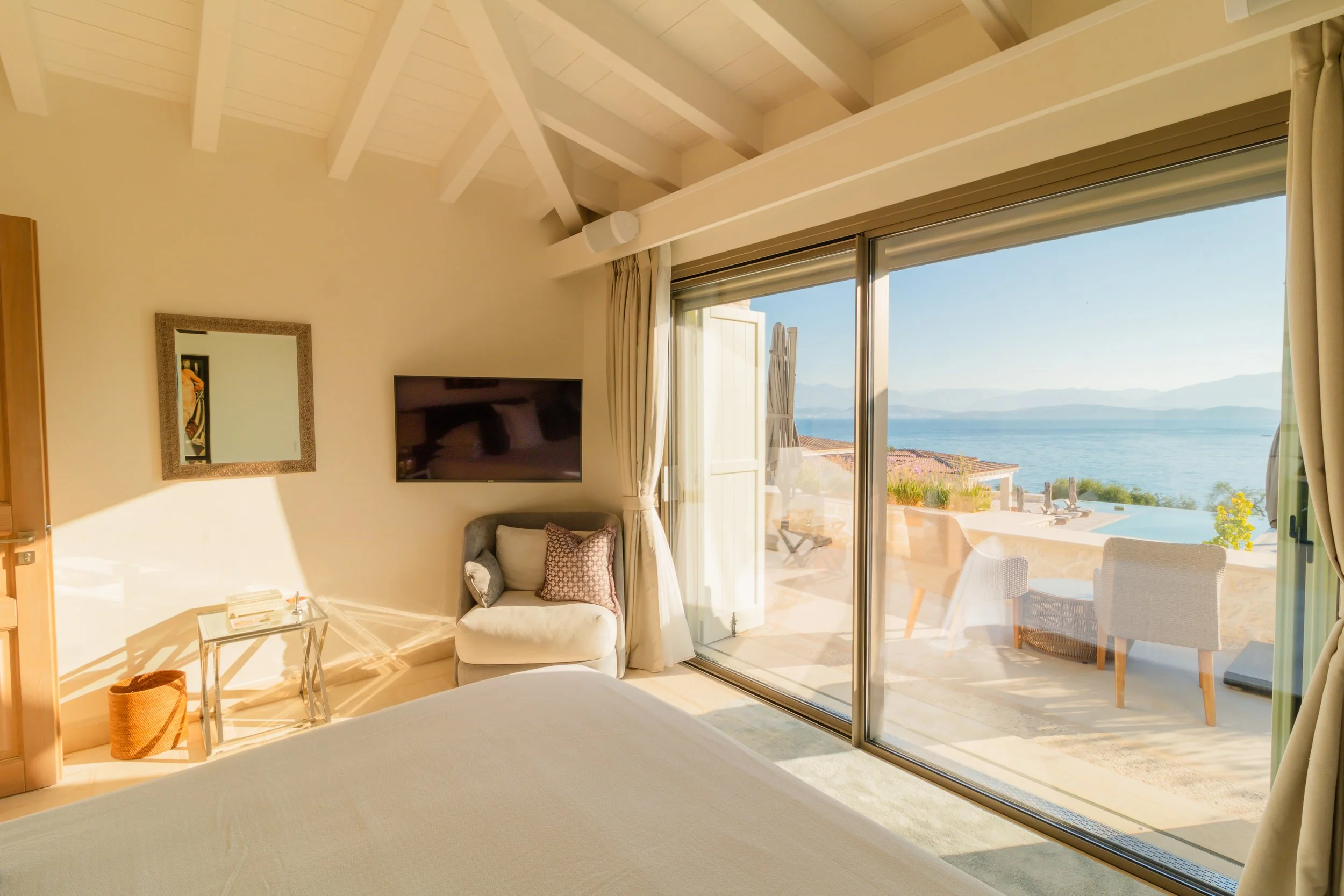 A bedroom with a large sliding glass door leading to a balcony overlooking a body of water and mountains in the distance, with outdoor seating and a pool.