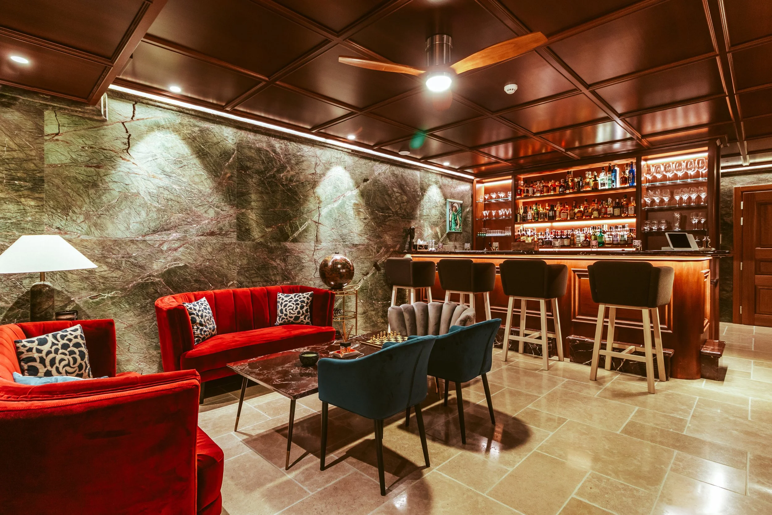 Interior of a sophisticated bar with a marble wall, red velvet couches, a modern bar counter with high chairs, and warm lighting.