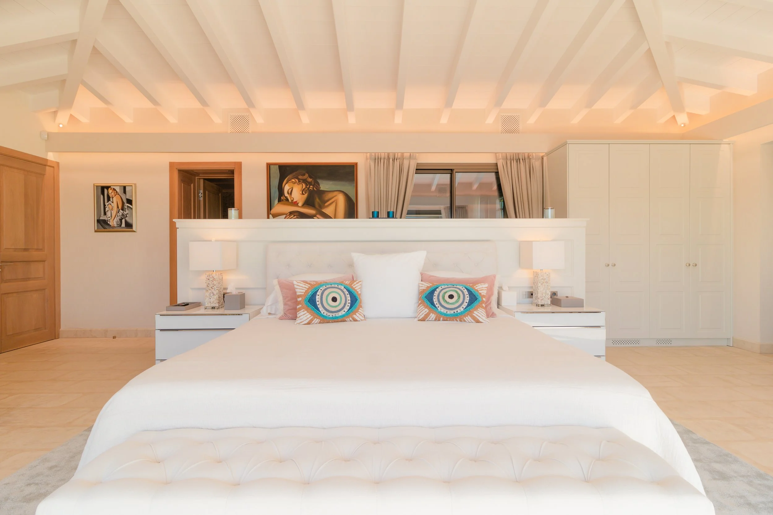 A bedroom with a white bed, colorful pillows, bedside lamps, artwork on the wall, and a storage cabinet.