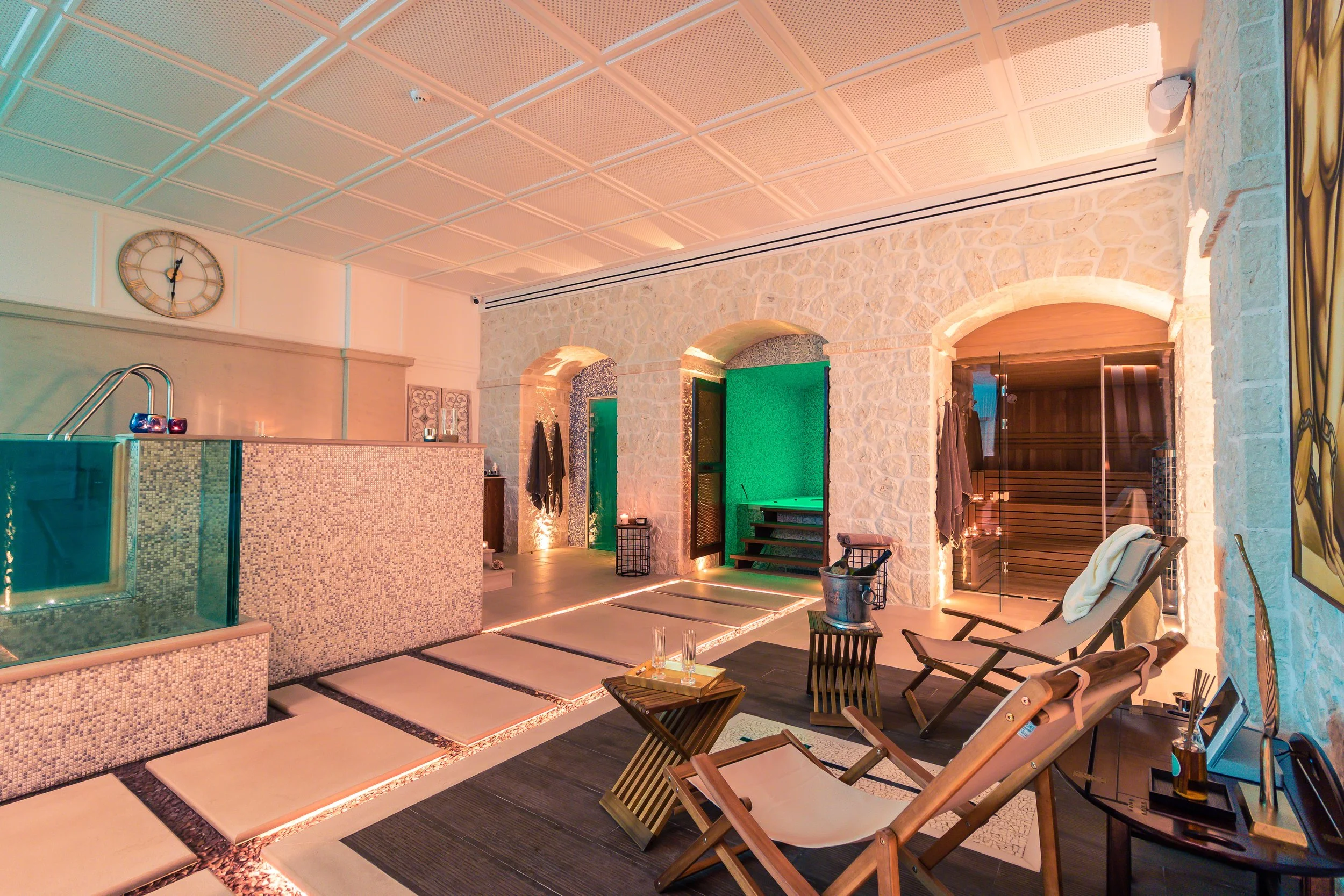 Spa room with lounge chairs, a small table, stone walls, sank, and a sauna room with glass door.