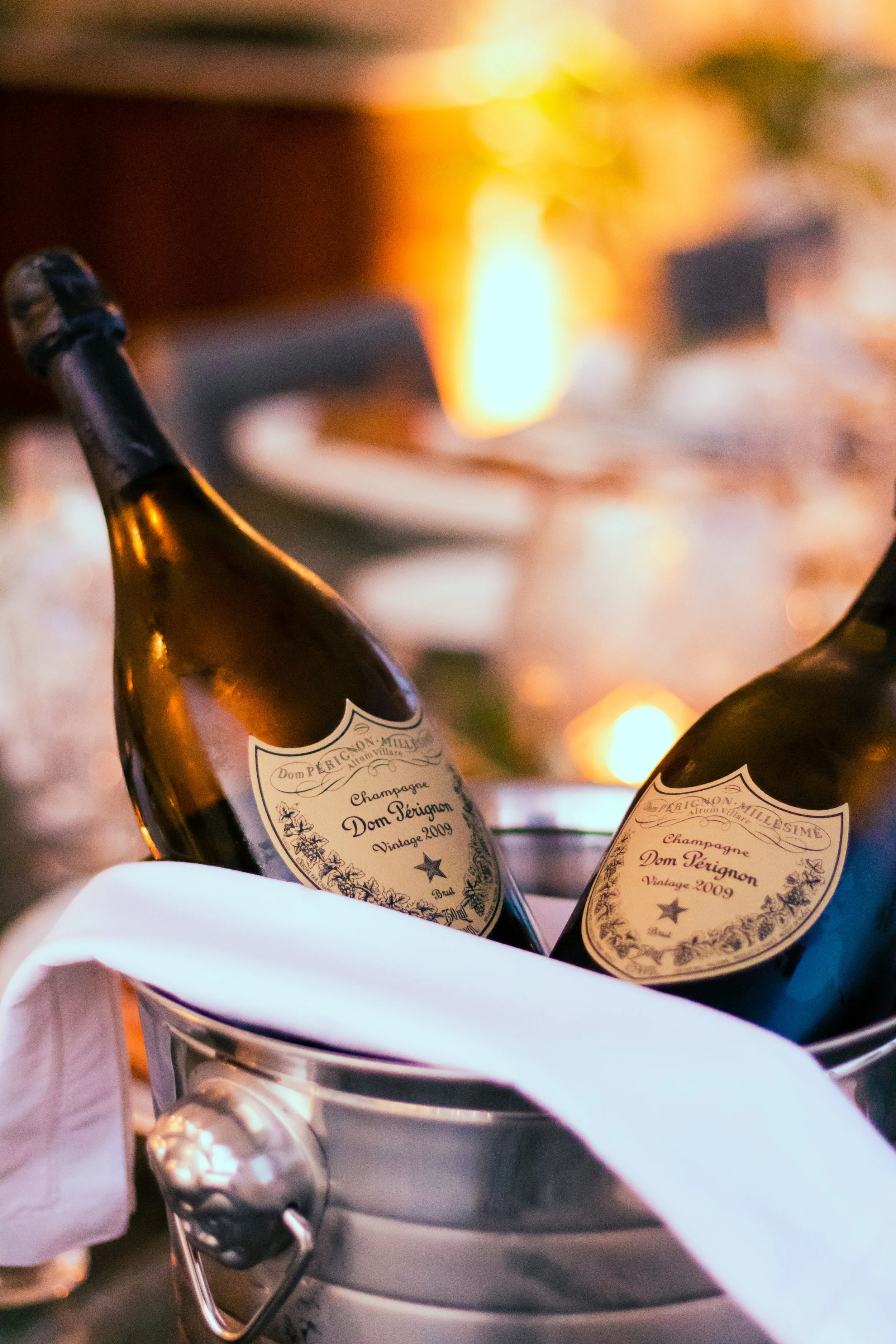 Two bottles of Dom Pérignon vintage 2009 champagne in an ice bucket with a white cloth, set against a warm, blurred background with soft lighting.