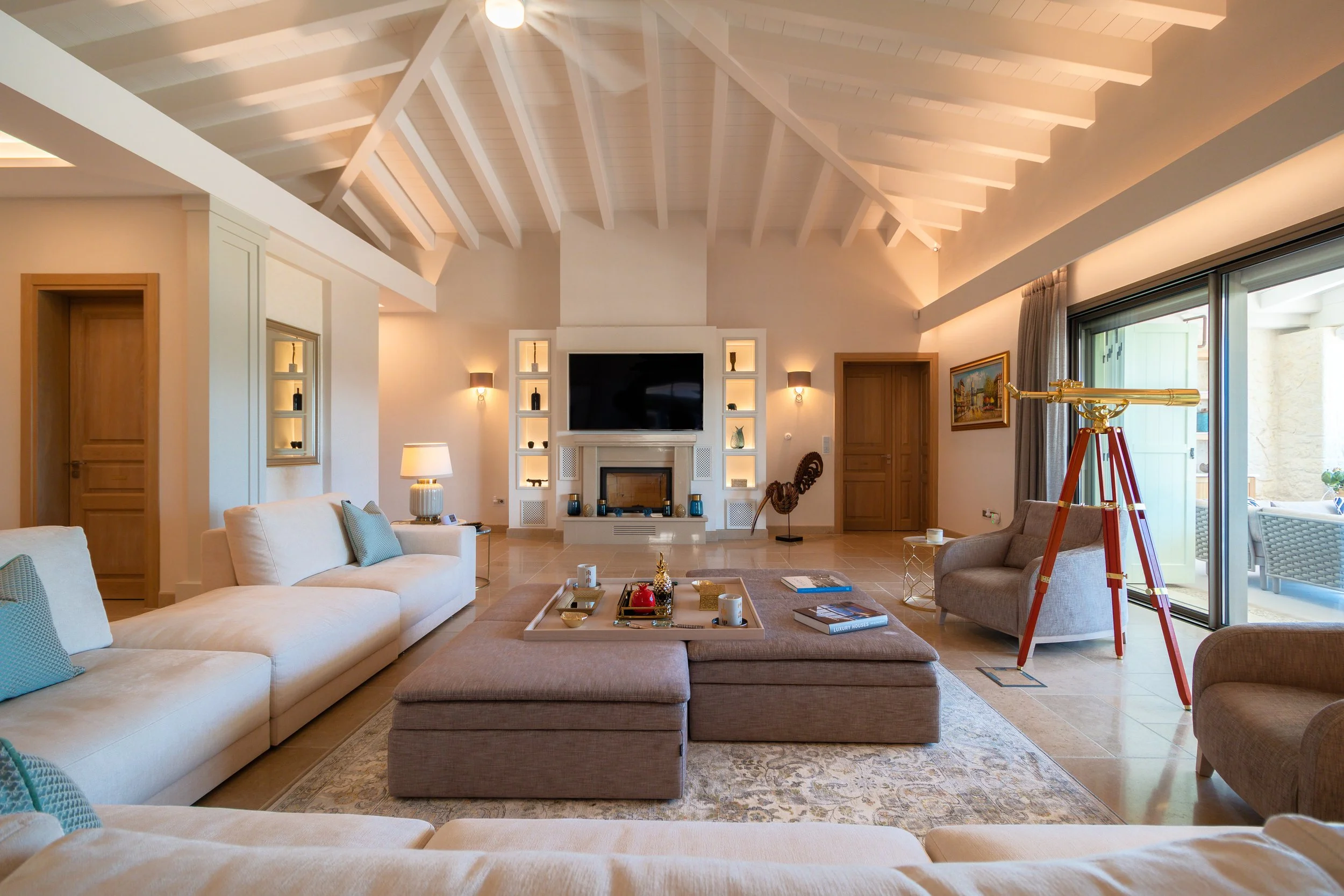Spacious living room with white vaulted ceiling and exposed beams, beige tiled floor, cream-colored sofas, a marble coffee table, a fireplace with a television mounted above, built-in shelves, a telescope near sliding glass doors, armchairs, and deco